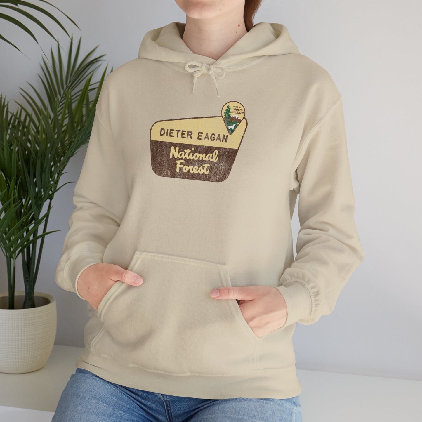Female model wearing a Sand colored Soft Unisex Hoodie Heavy Blend Hooded Sweatshirt featuring the logo graphic of the Dieter Eagan National Forest inspired by the TV show Severance in a vintage distressed ink-stamped worn-in style.