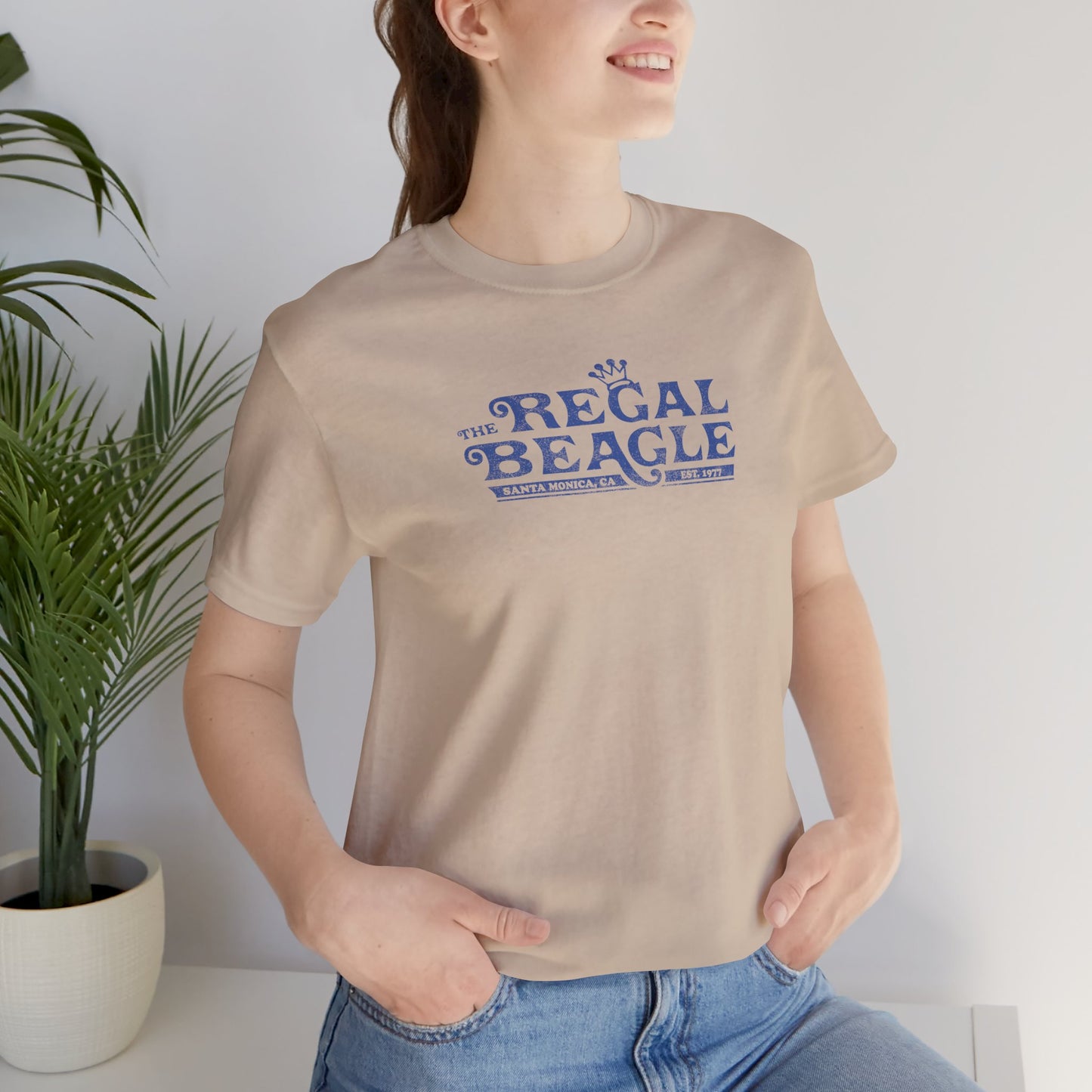 Female model wearing a tan colored soft Bella+Canvas 3001 short-sleeve t-shirt featuring the Regal Beagle logo from the TV Show Threes Company in a vintage distressed style.