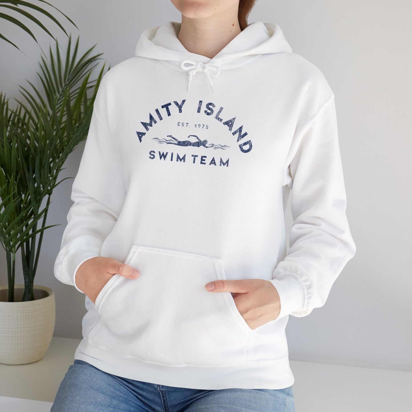Female model wearing a white colored Soft Unisex Hoodie Heavy Blend Hooded Sweatshirt featuring an Amity Island Swim Team logo graphic, designed by ScriptedBrand, and inspired by the movie Jaws in a vintage distressed style.