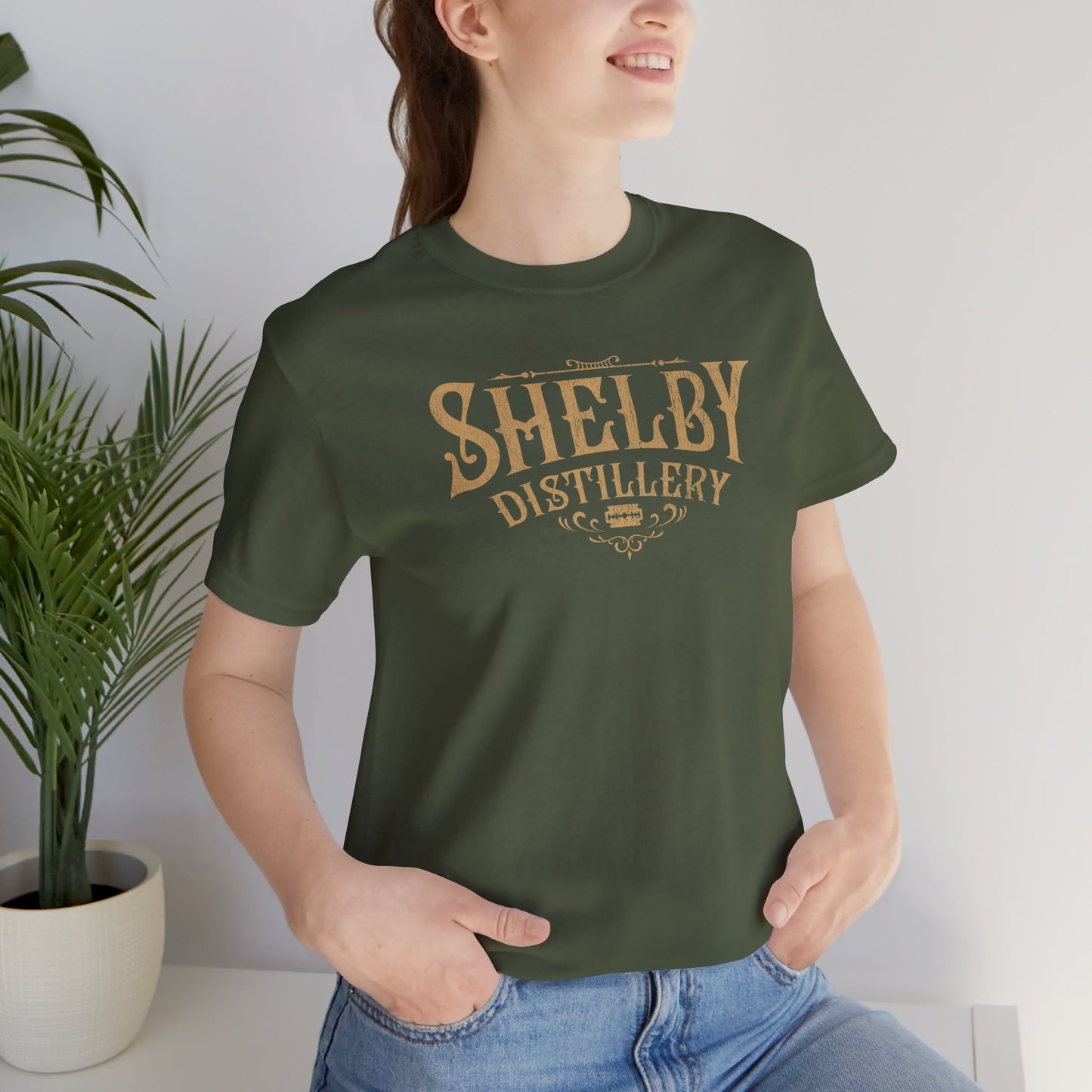 Female model wearing a military green Soft Bella+Canvas 3001 short-sleeve t-shirt featuring the Shelby Company Limited Distillery Logo Graphic inspired by the TV show and movie Peaky Blinders in a vintage distressed style, sold by ScriptedBrand.