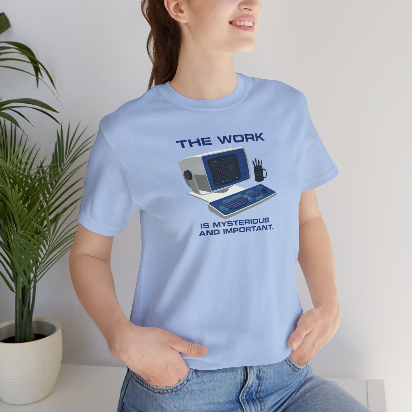 Female model wearing a baby blue Soft Bella+Canvas shirtsleeve t-shirt featuring the retro computer workstation from the Apple TV show Severance, and includes the text The Work is Mysterious and Important. Graphic is designed and illustrated by ScriptedBrand.