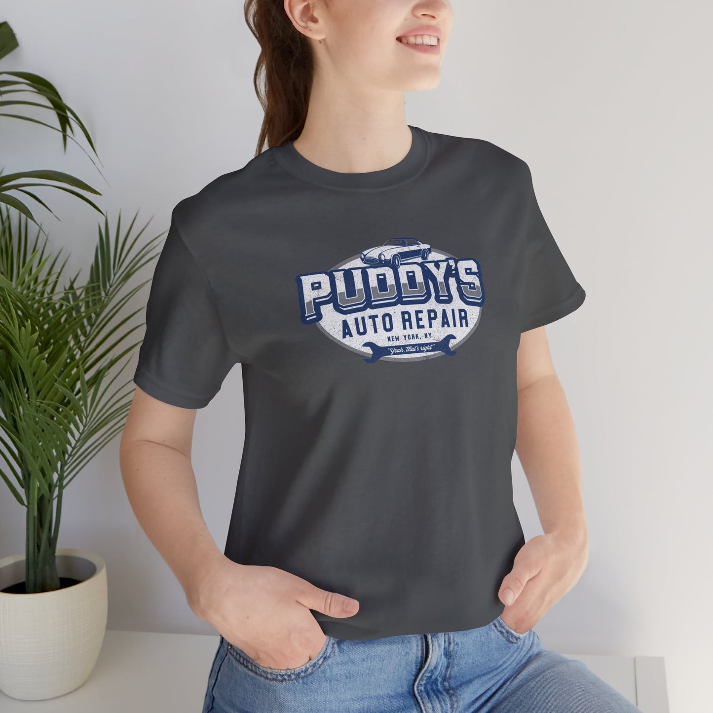 Female model wearing an asphalt gray soft short-sleeve t-shirt featuring a Puddy's Auto Repair graphic designed by ScriptedBrand inspired by the TV show Seinfeld in a vintage distressed ink-stamped worn-in style.