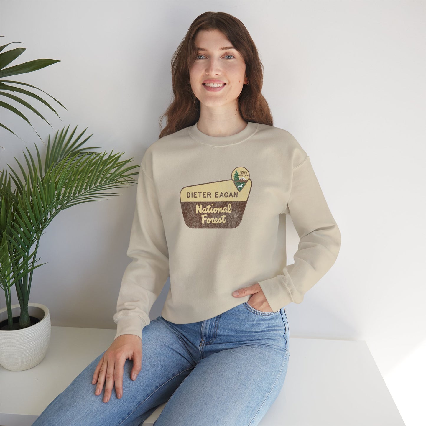 Female model wearing a sand tan Soft Unisex Gildan 18000 sweatshirt featuring a logo graphic of the Dieter Eagan National Forest sign designed by ScriptedBrand, inspired by the TV show Severance in a vintage distressed style.