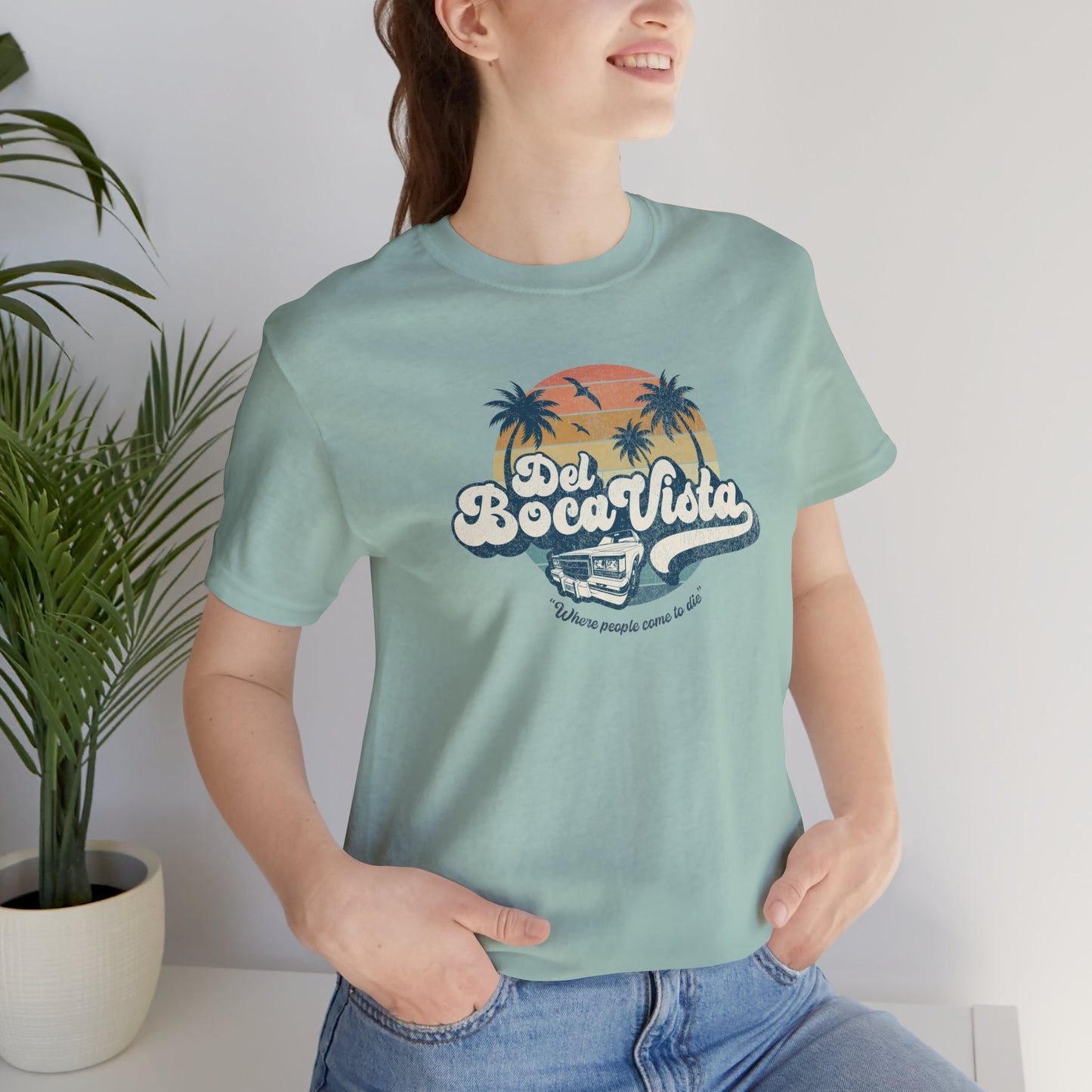 Female model wearing a dusty blue Soft Bella+Canvas 3001 short-sleeve t-shirt featuring a Del Boca Vista logo graphic (uniquely designed by ScriptedBrand), including the phrase Where People Come To Die, inspired by the TV show Seinfeld, in a vintage distressed style.