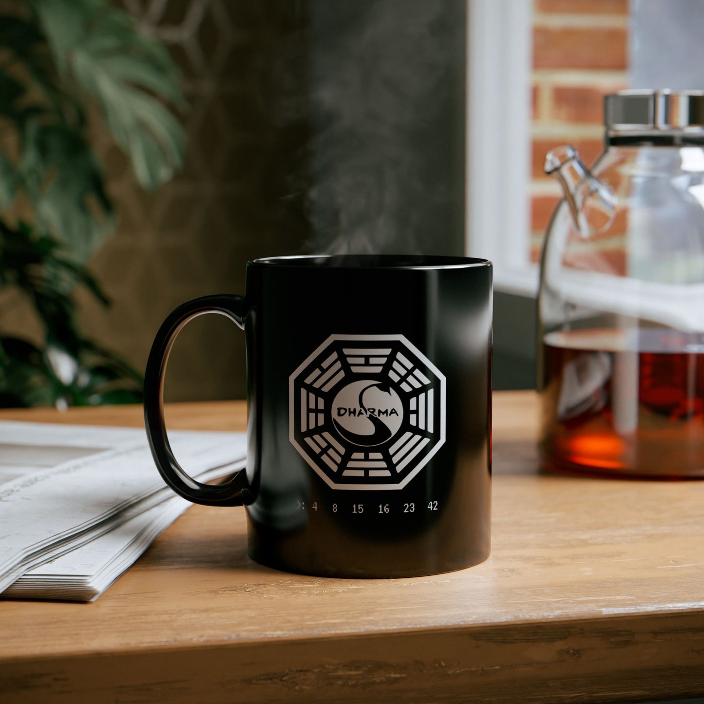 Black Ceramic Mug on a table with tea featuring the Dharma Initiative swan station logo from the TV show LOST, including the Hatch numbers 4 8 15 16 23 42. Graphic is printed white on both sides.