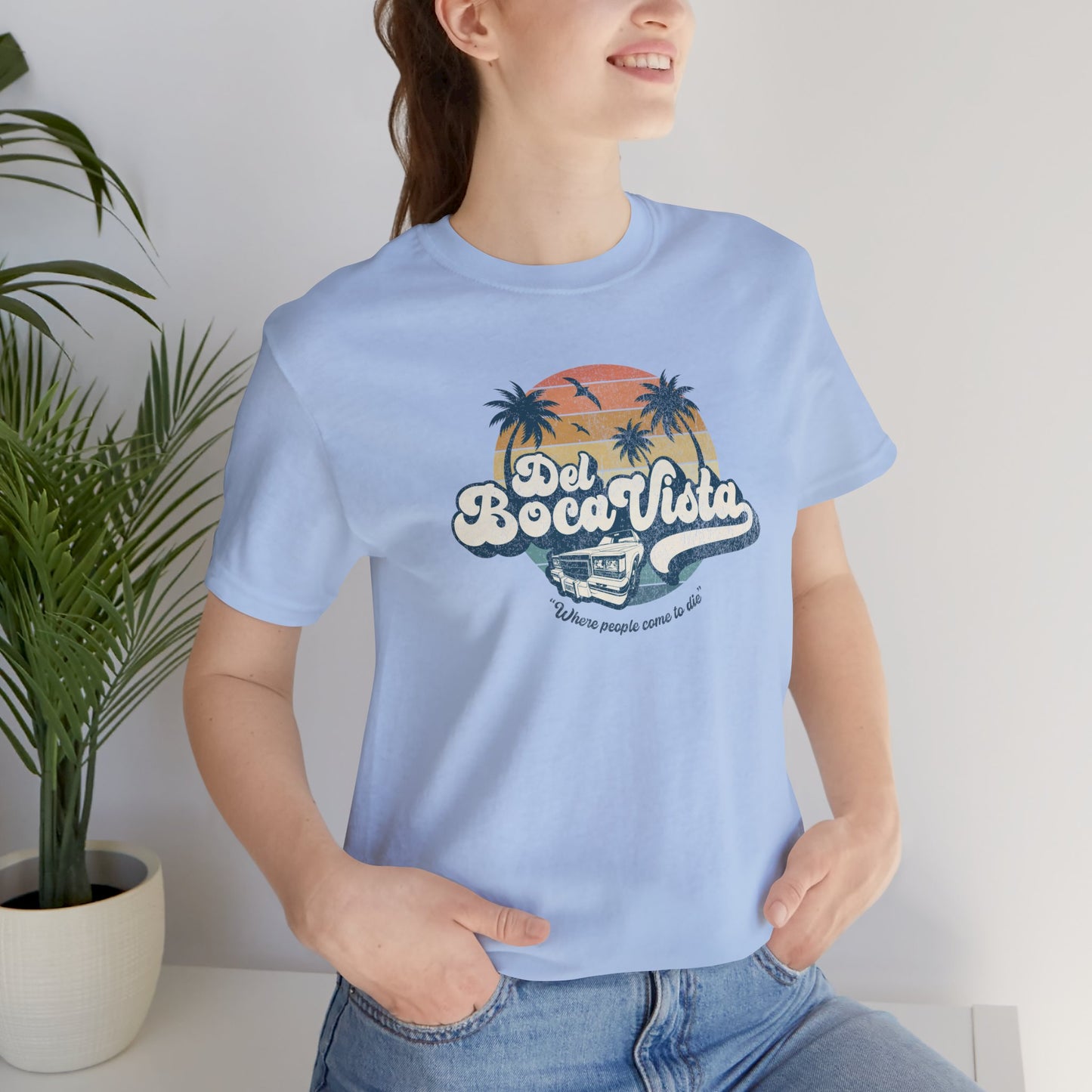 Female model wearing a baby blue Soft Bella+Canvas 3001 short-sleeve t-shirt featuring a Del Boca Vista logo graphic (uniquely designed by ScriptedBrand), including the phrase Where People Come To Die, inspired by the TV show Seinfeld, in a vintage distressed style.