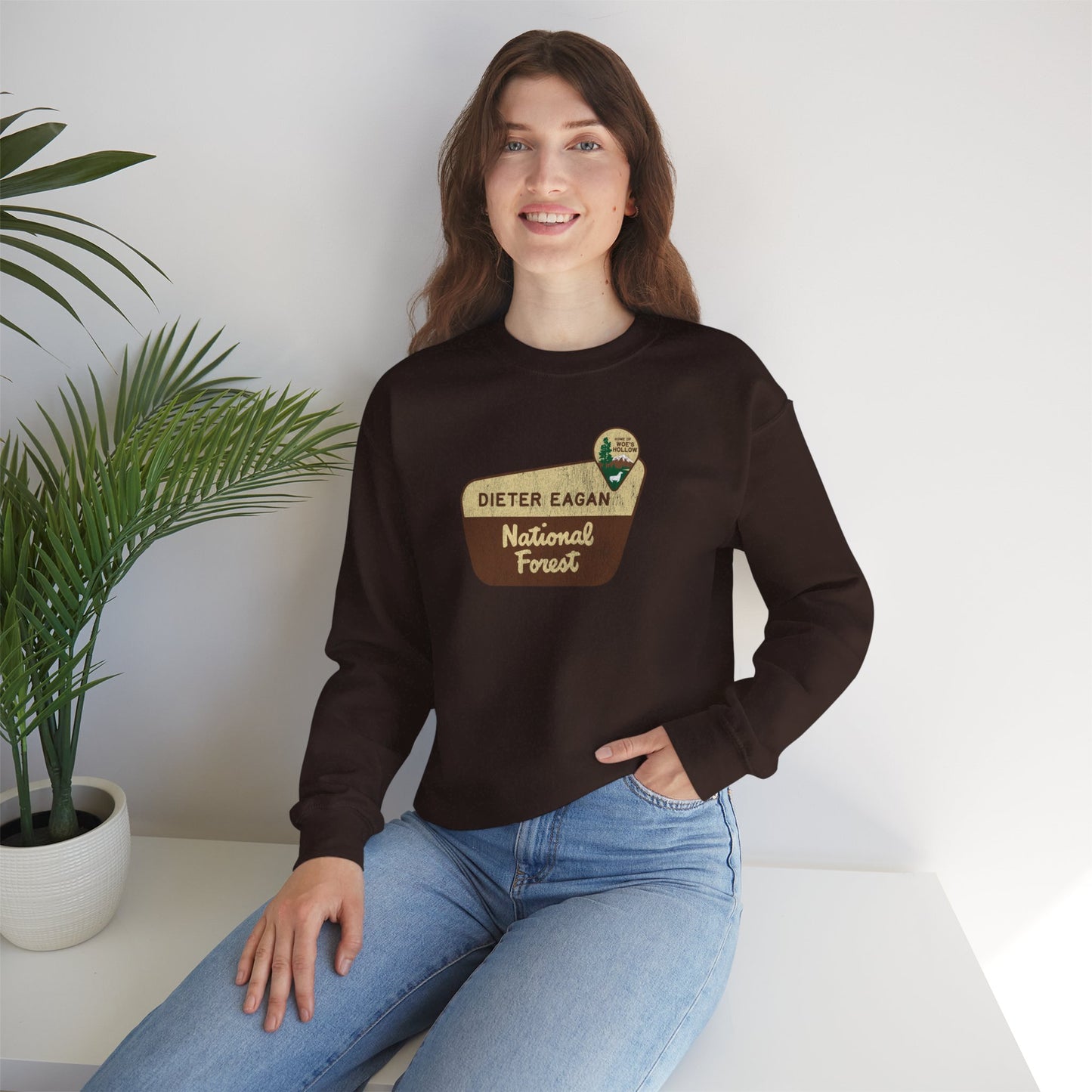 Female model wearing a dark chocolate brown Soft Unisex Gildan 18000 sweatshirt featuring a logo graphic of the Dieter Eagan National Forest sign designed by ScriptedBrand, inspired by the TV show Severance in a vintage distressed style.
