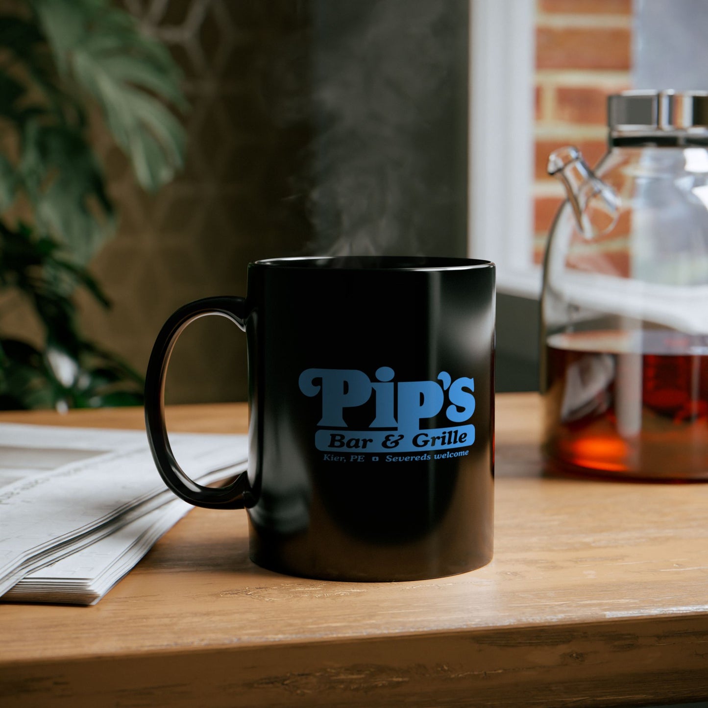 Mockup on a table of a 11 oz Black Ceramic Mug featuring the Pips Bar and Grill logo from the Apple TV show Severance printed on both sides in Lumon Industries blue, sold by ScriptedBrand. Mug is filled with hot tea.