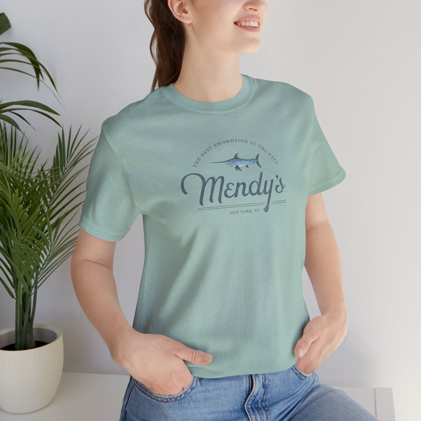 Female model wearing a dusty blue colored soft unisex Bella+Canvas 3001 short-sleeve t-shirt featuring a unique Mendy's Restaurant logo graphic, designed by ScriptedBrand in a vintage distressed style, and inspired by the TV show Seinfeld.