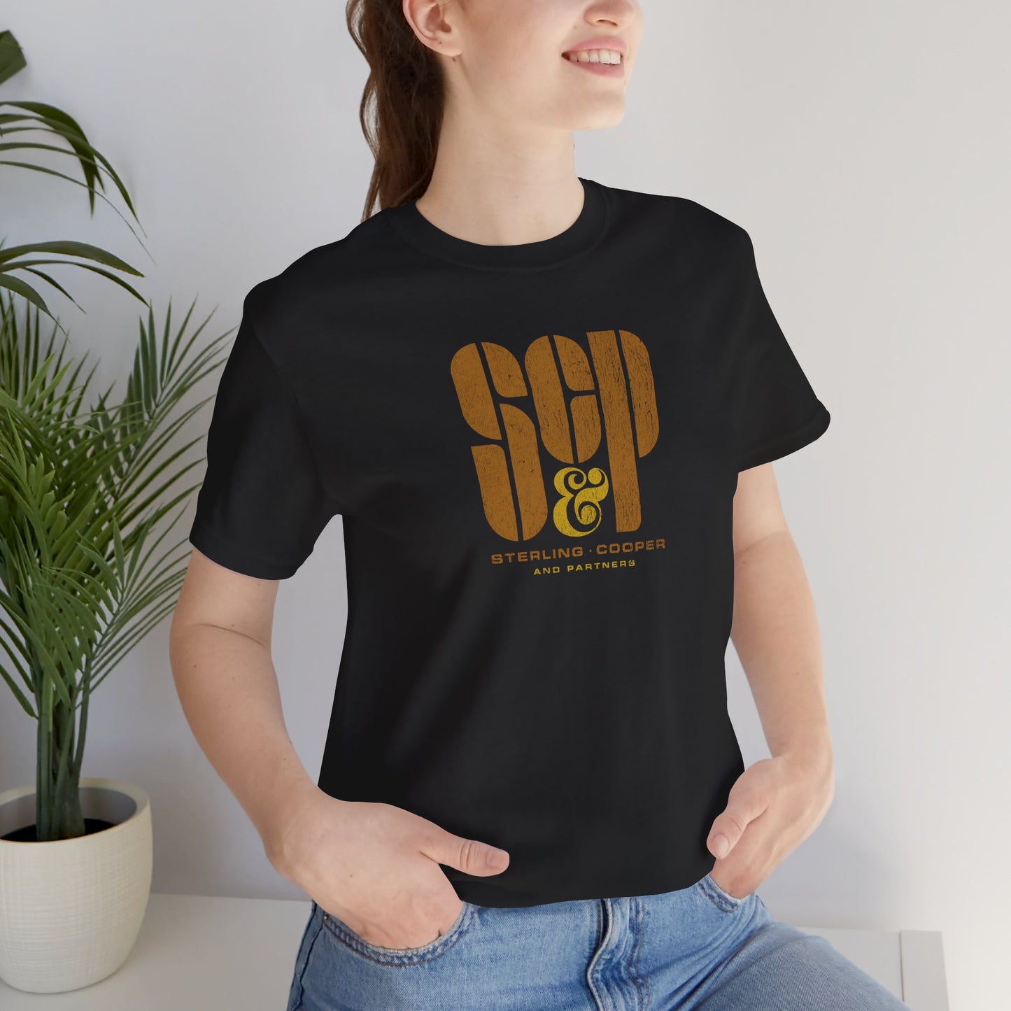 Female model wearing a black colored soft unisex Bella+Canvas 3001 short-sleeve t-shirt featuring the Sterling Cooper & Partners logo graphic from the TV Show Mad Men in a vintage distressed style, sold by ScriptedBrand.