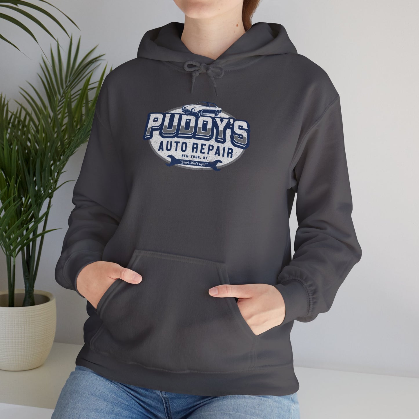 Female model wearing a charcoal gray soft Unisex Hoodie Heavy Blend Hooded Sweatshirt featuring a Puddy's Auto Repair graphic designed by ScriptedBrand inspired by the TV show Seinfeld in a vintage distressed style.
