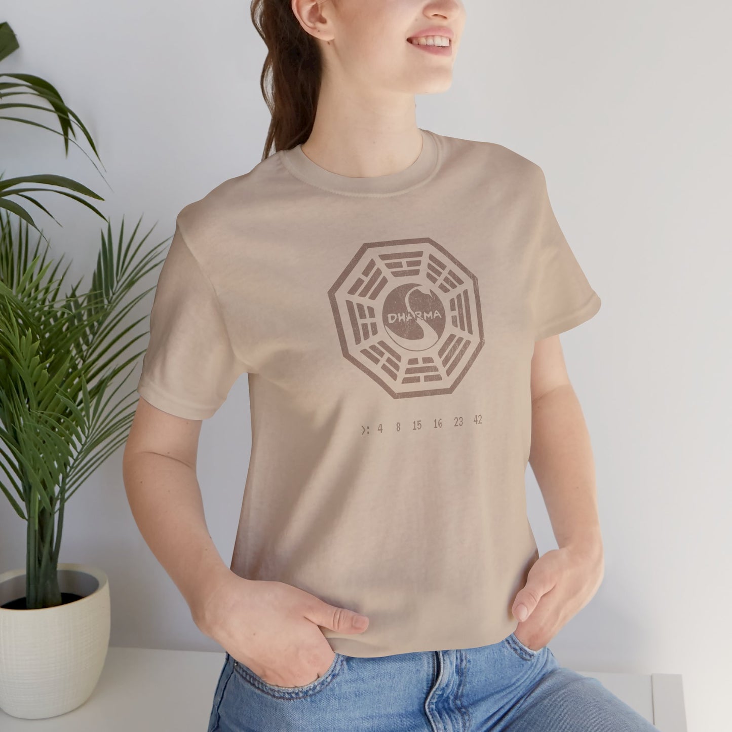 Female model wearing a Tan short-sleeve t-shirt featuring the Dharma Initiative swan station logo graphic from the TV show LOST in a vintage distressed style.