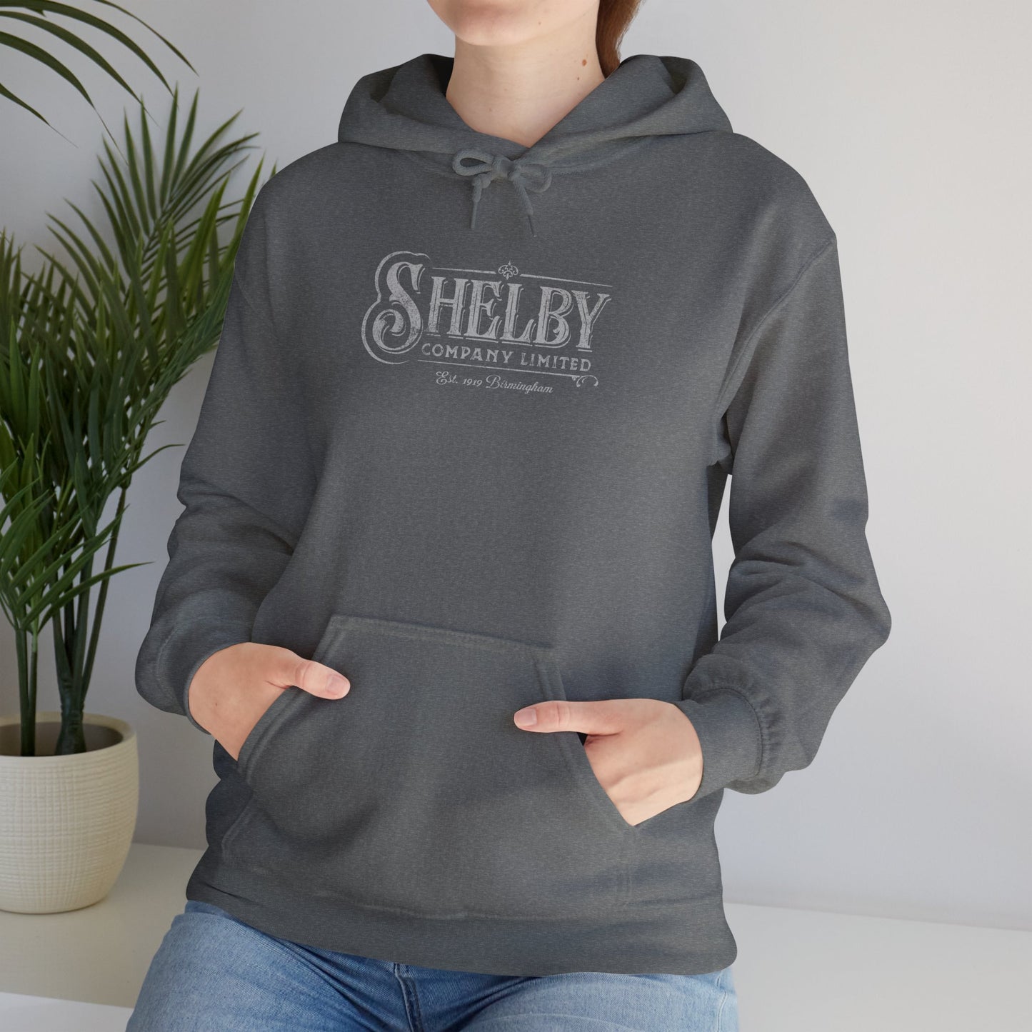 Female model wearing a dark heather gray colored Soft Unisex Hoodie Heavy Blend Hooded Sweatshirt featuring the fictional logo Shelby Company Limited from the TV series Peaky Blinders in a vintage distressed style.