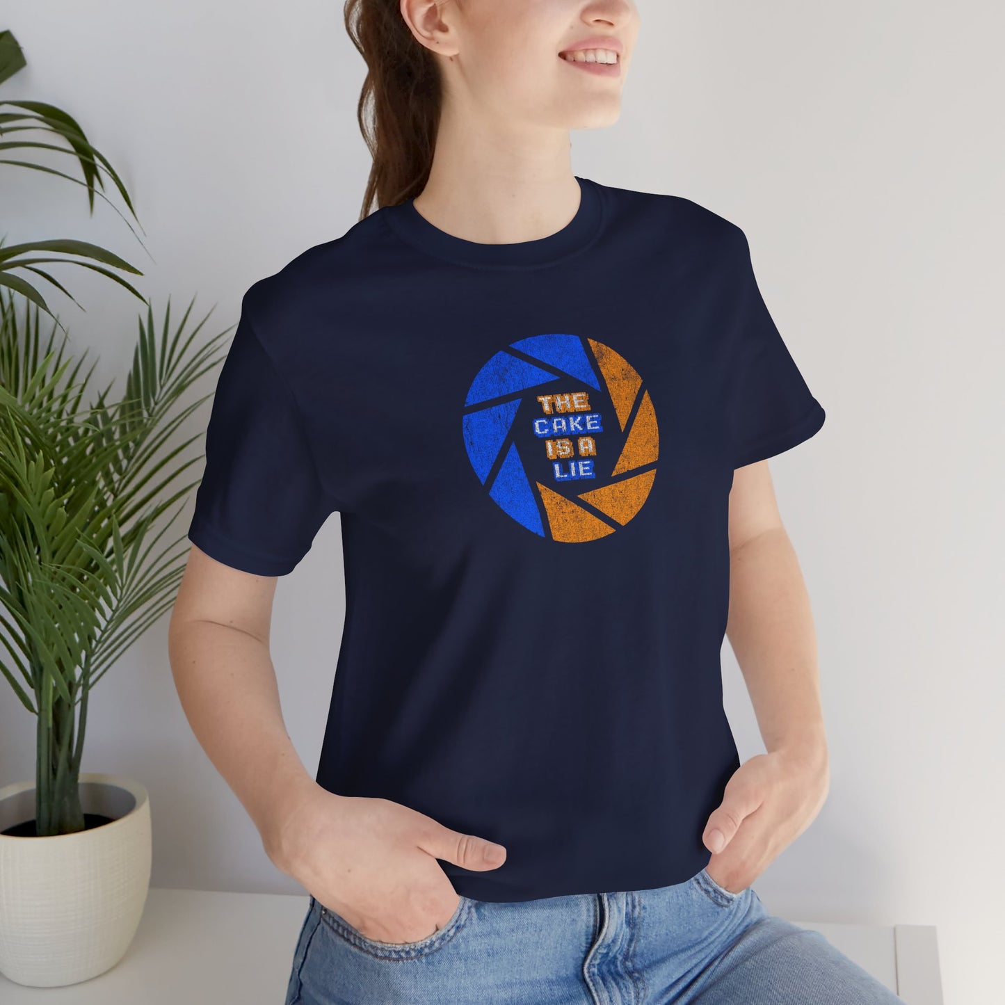 Female model wearing a navy blue colored soft Bella+Canvas 3001 short-sleeve t-shirt featuring the phrase The Cake Is a Lie, along with the Aperture Laboratories shutter logo from the video game Portal and Portal 2 in a vintage distressed style, sold by ScriptedBrand.