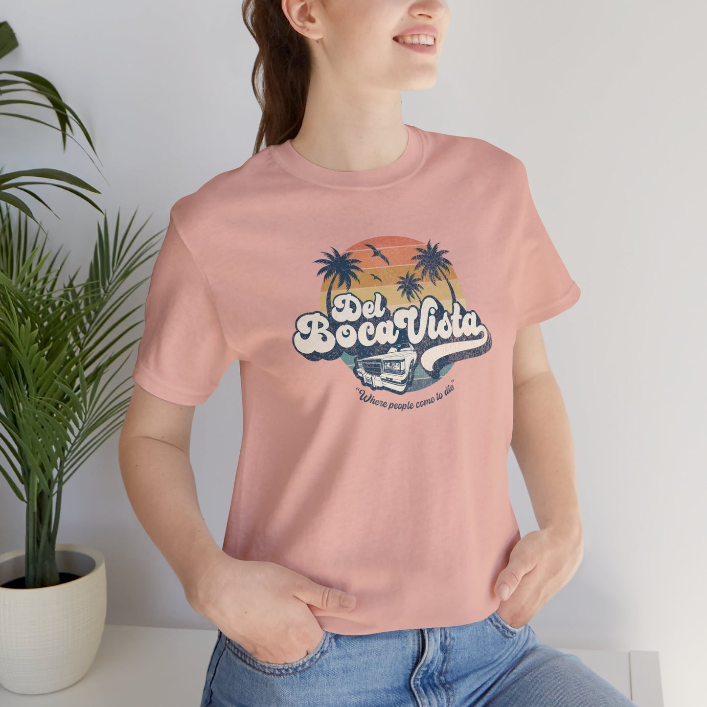 Female model wearing a peach Soft Bella+Canvas 3001 short-sleeve t-shirt featuring a Del Boca Vista logo graphic (uniquely designed by ScriptedBrand), including the phrase Where People Come To Die, inspired by the TV show Seinfeld, in a vintage distressed style.