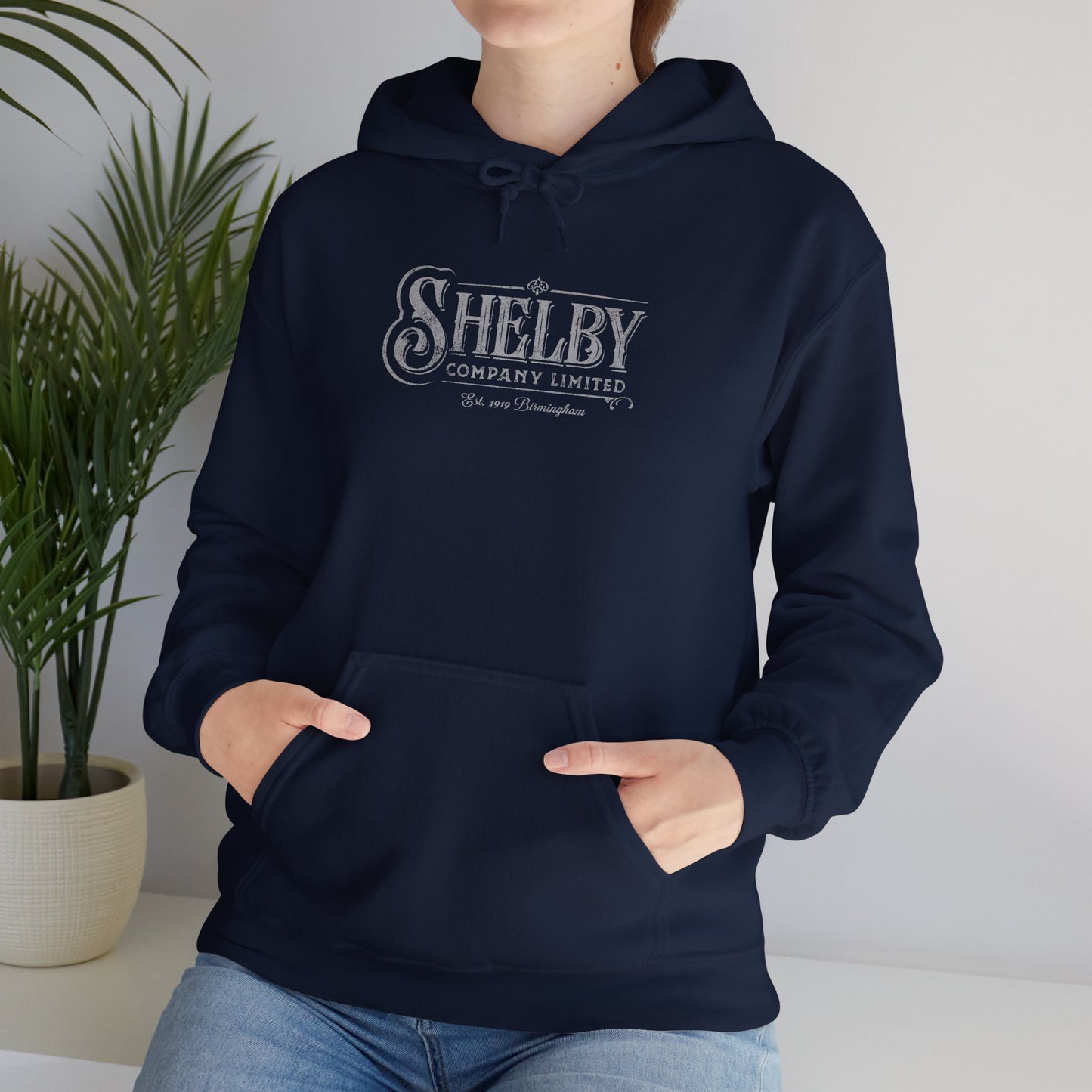 Female model wearing a navy blue colored Soft Unisex Hoodie Heavy Blend Hooded Sweatshirt featuring the fictional logo Shelby Company Limited from the TV series Peaky Blinders in a vintage distressed style.