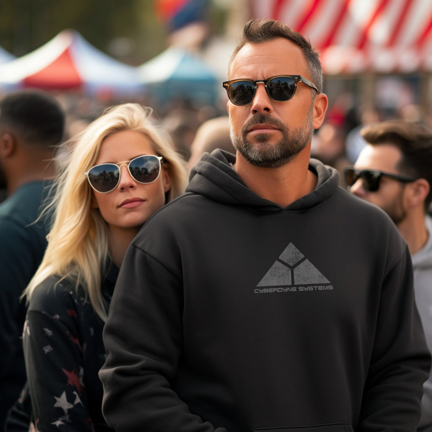 Man wearing a black hoodie featuring the fictional logo Skynet Cyberdyne Systems from the movie The Terminator printed on both sides in a vintage distressed style, surrounded by people at an outdoor event.