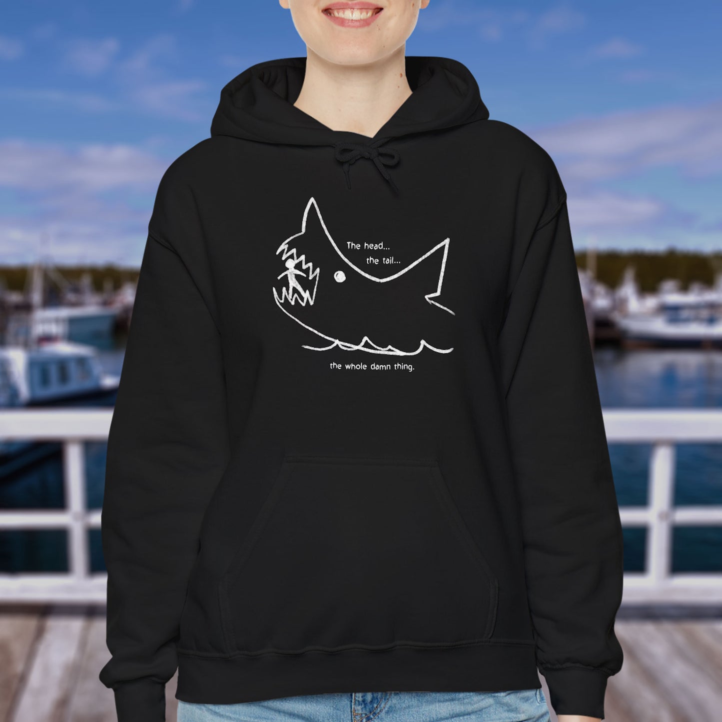 Female model wearing a black colored Soft Unisex Hoodie Heavy Blend Hooded Sweatshirt with the chalkboard drawing of a shark eating a person, drawn by Quint in the movie Jaws.
