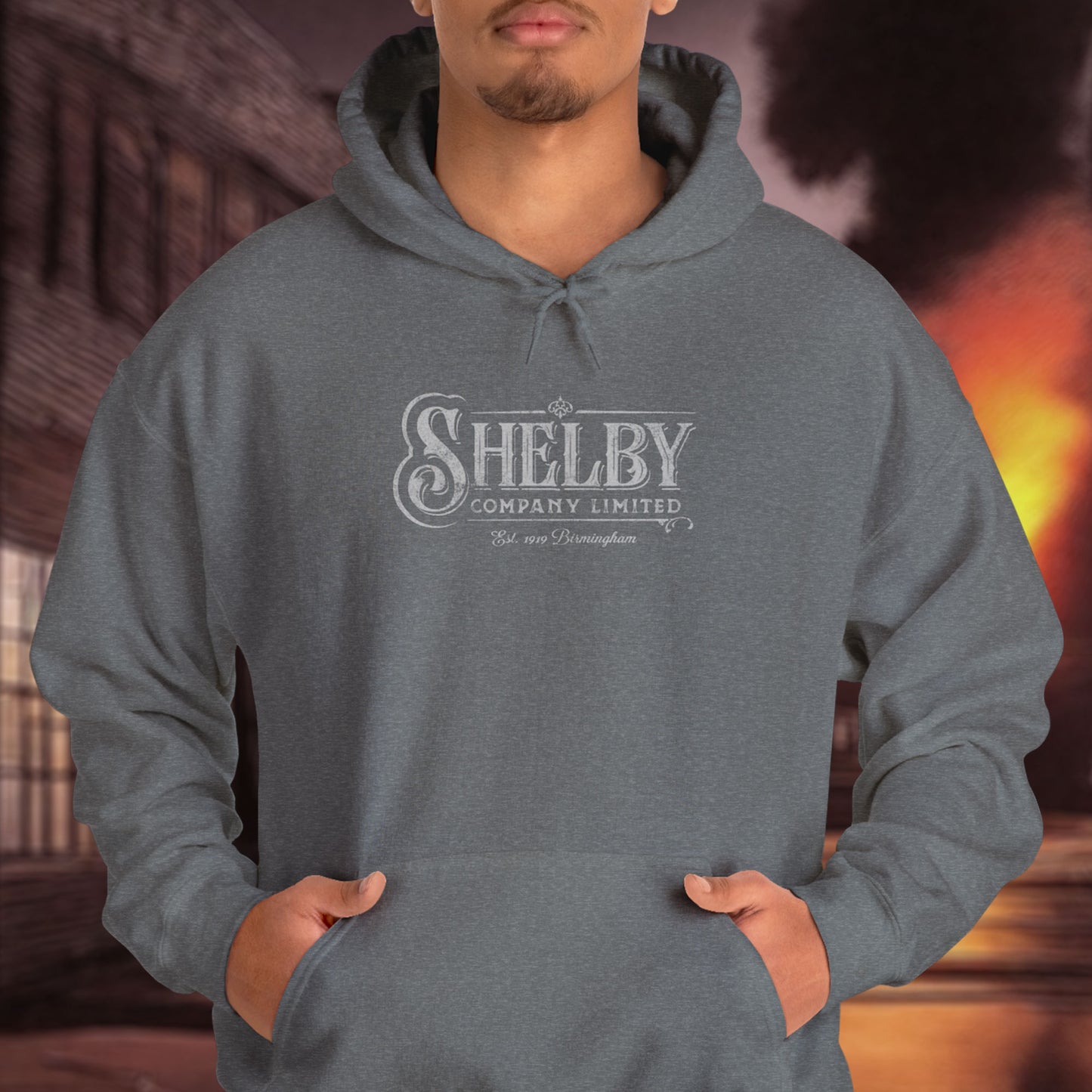 Male model wearing a dark heather gray colored Soft Unisex Hoodie Heavy Blend Hooded Sweatshirt featuring the fictional logo Shelby Company Limited from the TV series Peaky Blinders in a vintage distressed style.