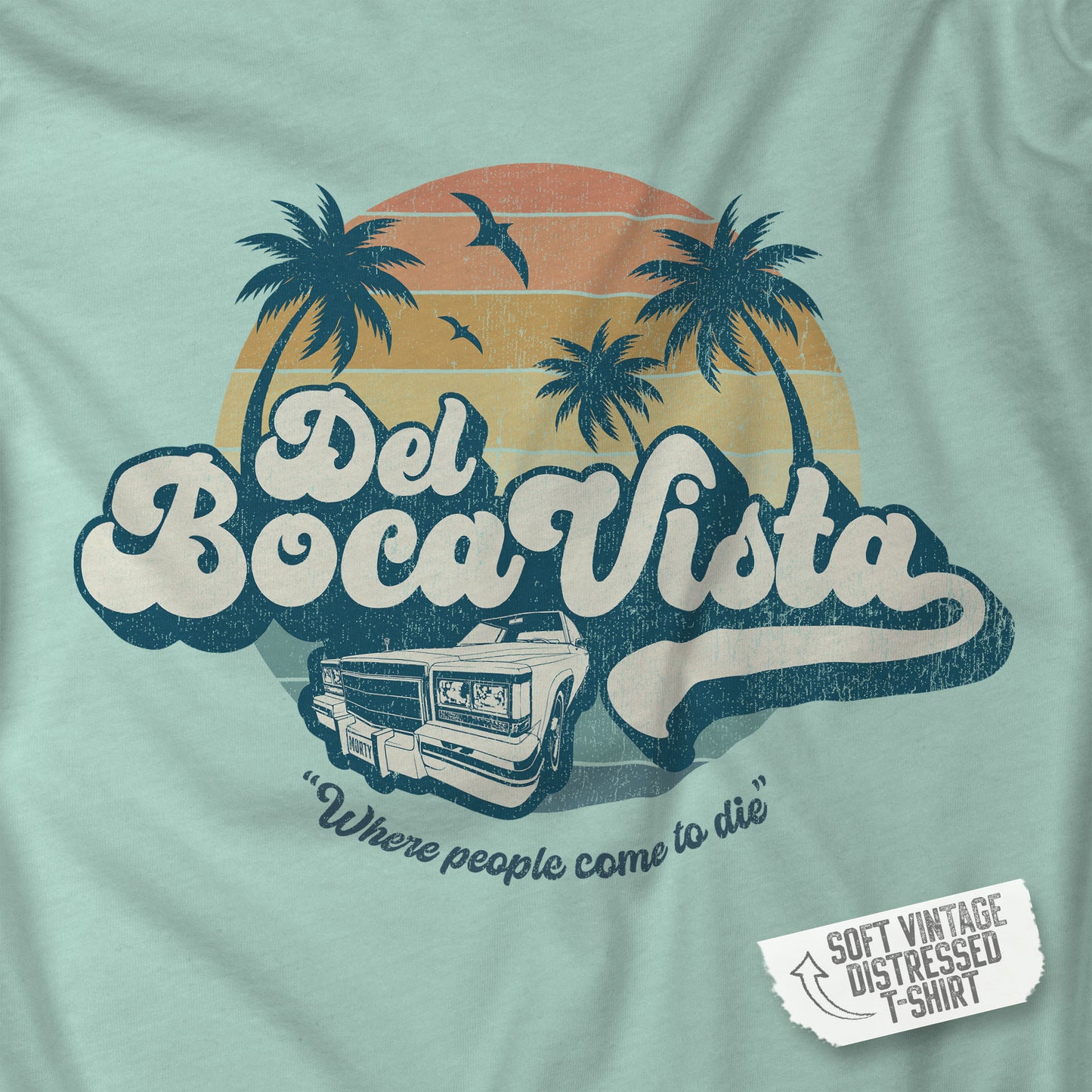 Closeup of a dusty blue Soft Bella+Canvas 3001 short-sleeve t-shirt featuring a Del Boca Vista logo graphic (uniquely designed by ScriptedBrand), including the phrase Where People Come To Die, inspired by the TV show Seinfeld, in a vintage distressed style.