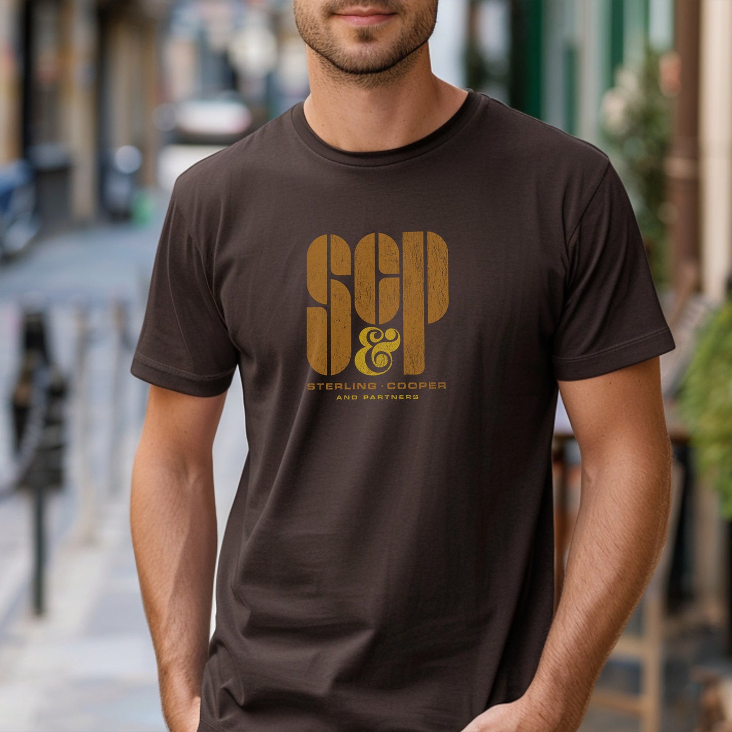 Male model wearing a brown colored soft unisex Bella+Canvas 3001 short-sleeve t-shirt featuring the Sterling Cooper & Partners logo graphic from the TV Show Mad Men in a vintage distressed style, sold by ScriptedBrand.