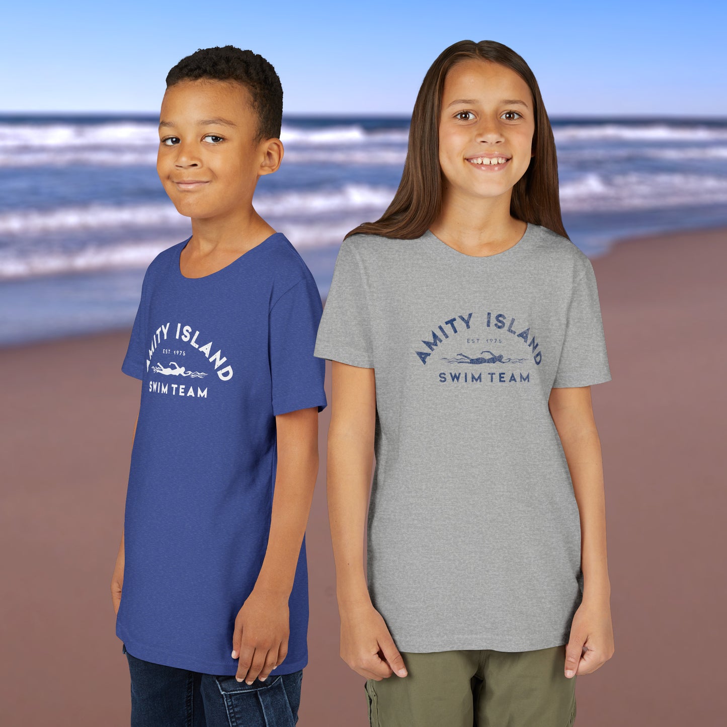 2 kid models wearing royal blue and heather grey colored Soft unisex Youth Sizes Bella+Canvas 3001Y short-sleeve t-shirts featuring an Amity Island Swim Team logo graphic, designed by ScriptedBrand, and inspired by the movie Jaws in a vintage distressed style.