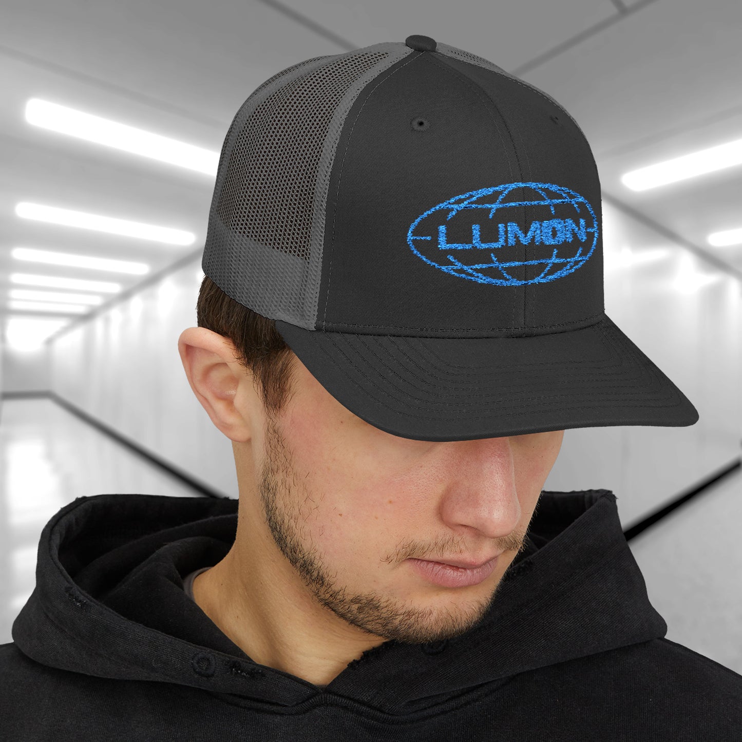 Male model wearing a black/charcoal gray Embroidered Snapback Trucker Cap featuring the blue logo of Lumon Industries from the Apple TV Show Severance.