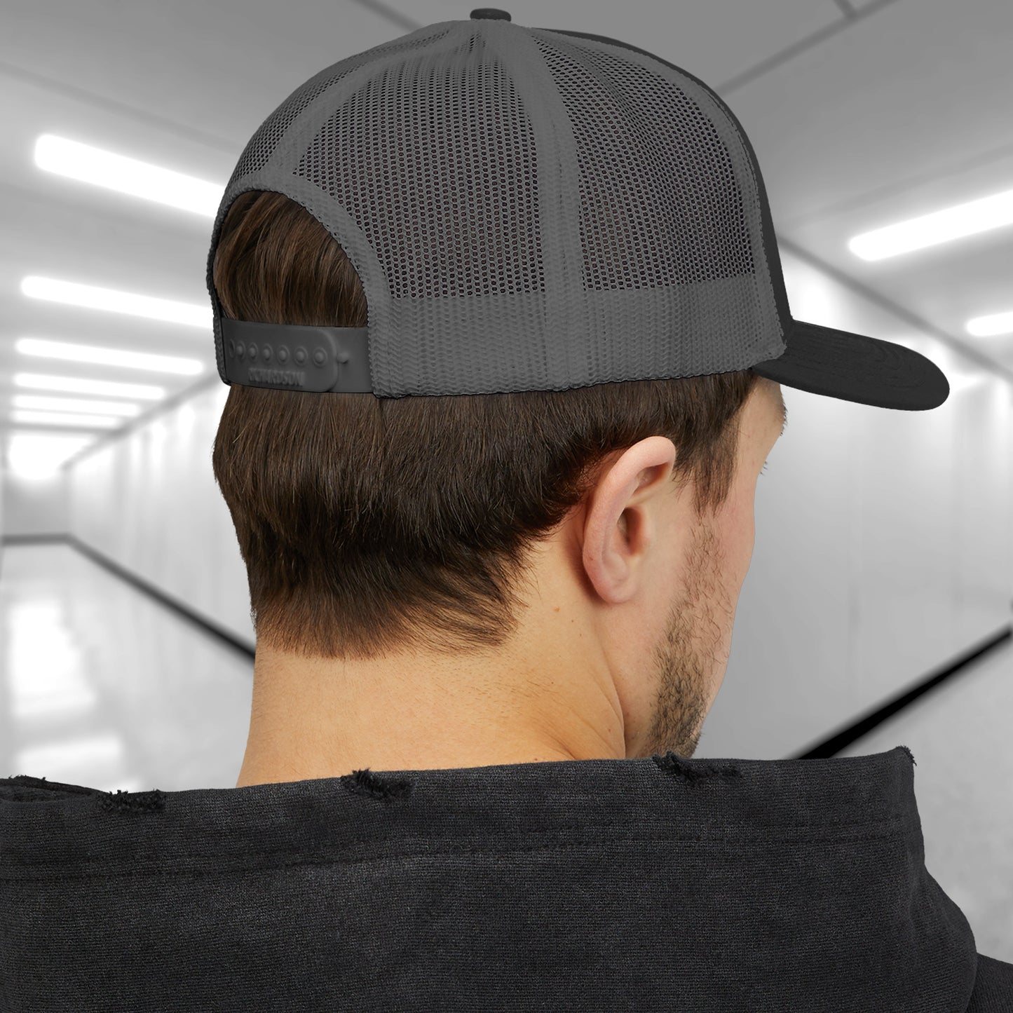 Back view of a male model wearing a black/charcoal gray Embroidered Snapback Trucker Cap featuring the blue logo of Lumon Industries from the Apple TV Show Severance.