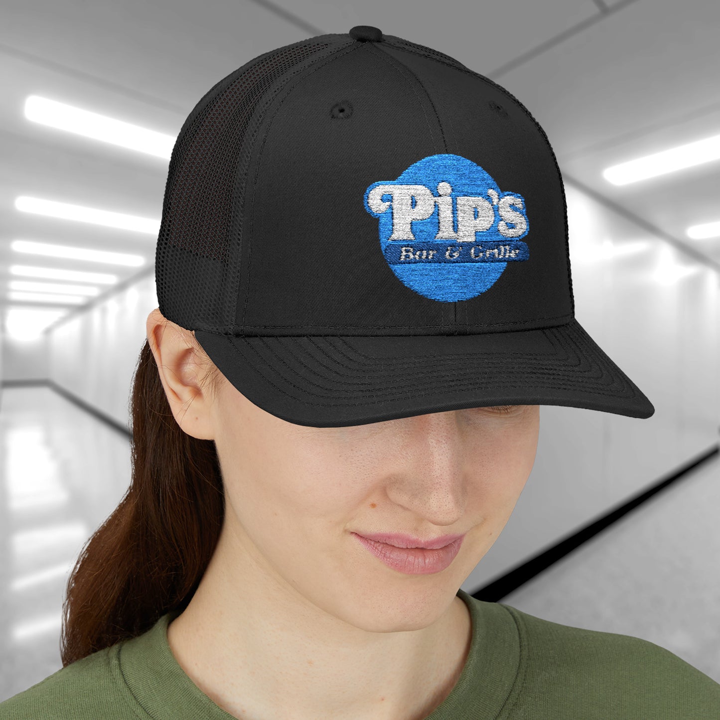 Female model wearing a black Snapback Trucker Cap featuring an embroidered Pip's Bar and Grill logo from the Apple TV show Severance.