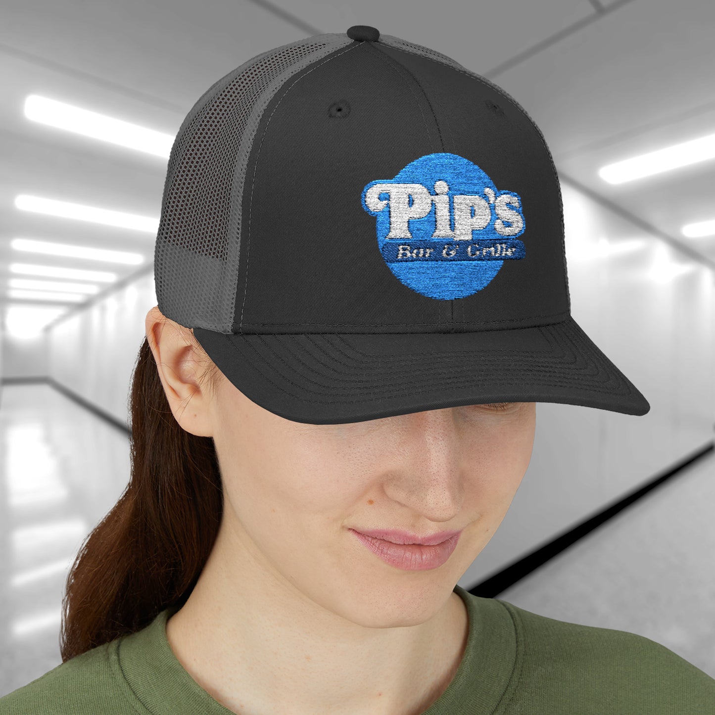 Female model wearing a black/charcoal Snapback Trucker Cap featuring an embroidered Pip's Bar and Grill logo from the Apple TV show Severance.