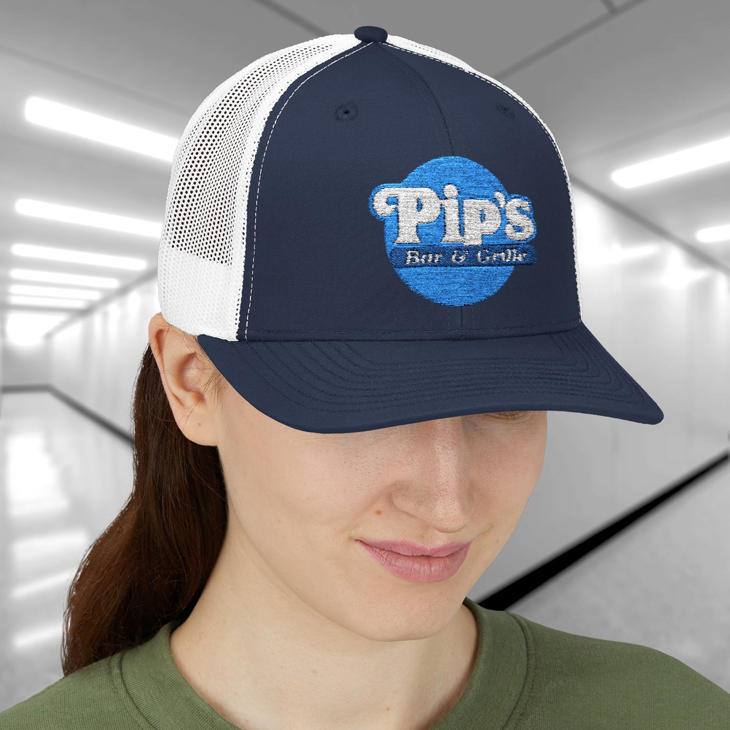 Female model wearing a navy/white Snapback Trucker Cap featuring an embroidered Pip's Bar and Grill logo from the Apple TV show Severance.