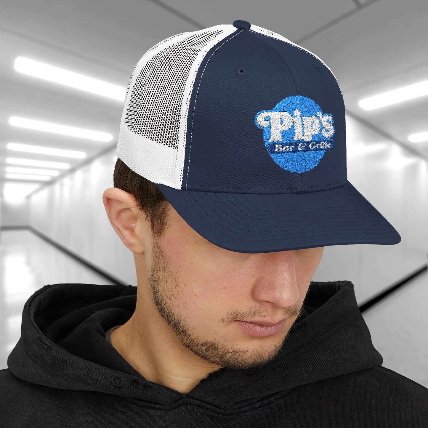 Male model wearing a navy/white Snapback Trucker Cap featuring an embroidered Pip's Bar and Grill logo from the Apple TV show Severance.