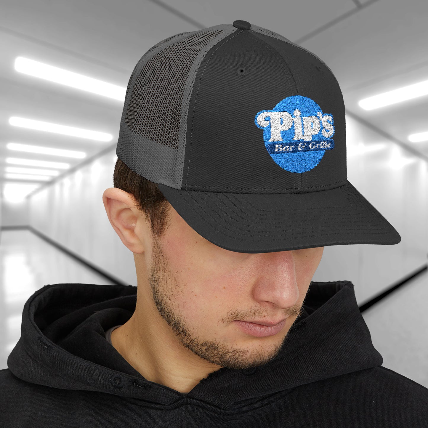 Male model wearing a black/charcoal Snapback Trucker Cap featuring an embroidered Pip's Bar and Grill logo from the Apple TV show Severance.