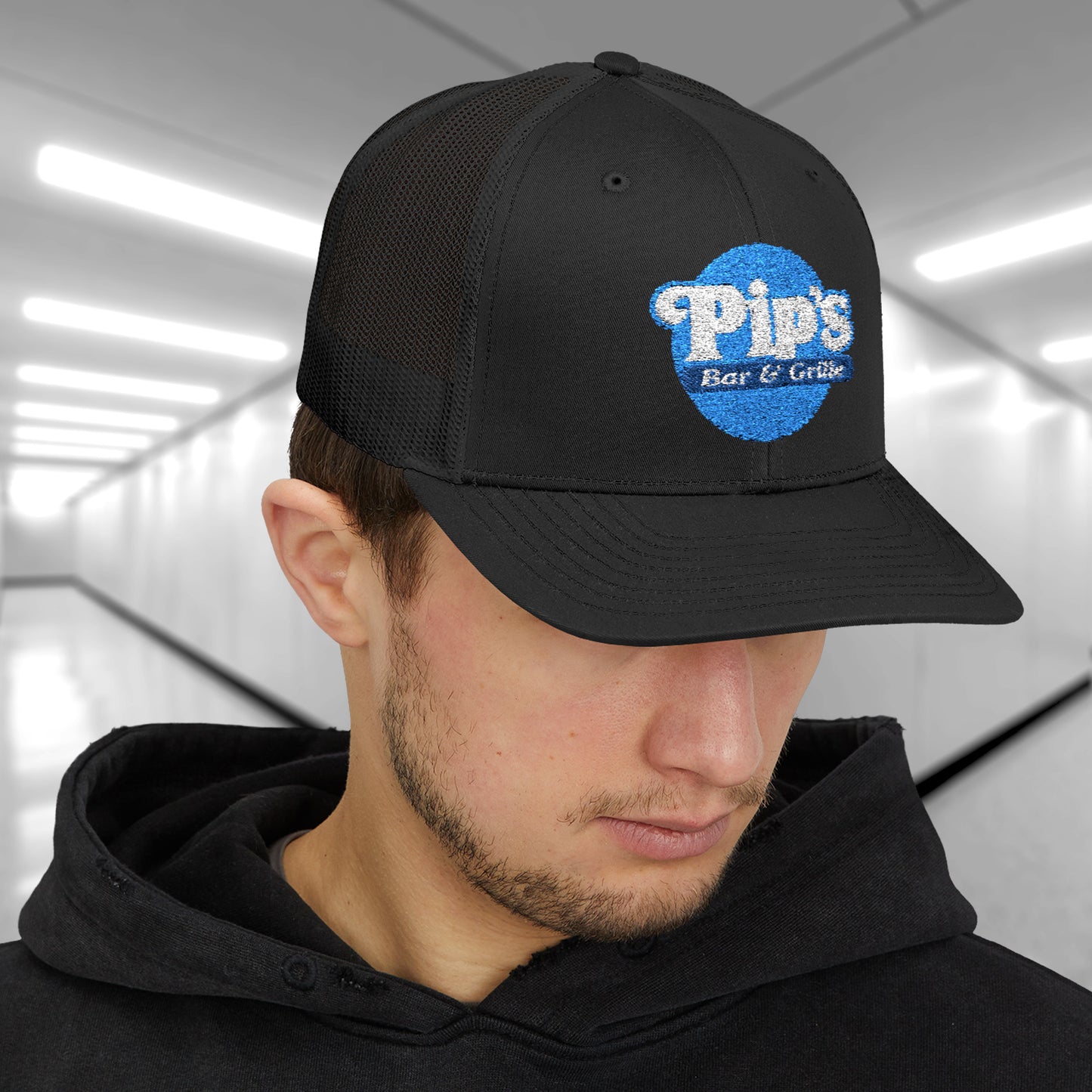 Male model wearing a black Snapback Trucker Cap featuring an embroidered Pip's Bar and Grill logo from the Apple TV show Severance.
