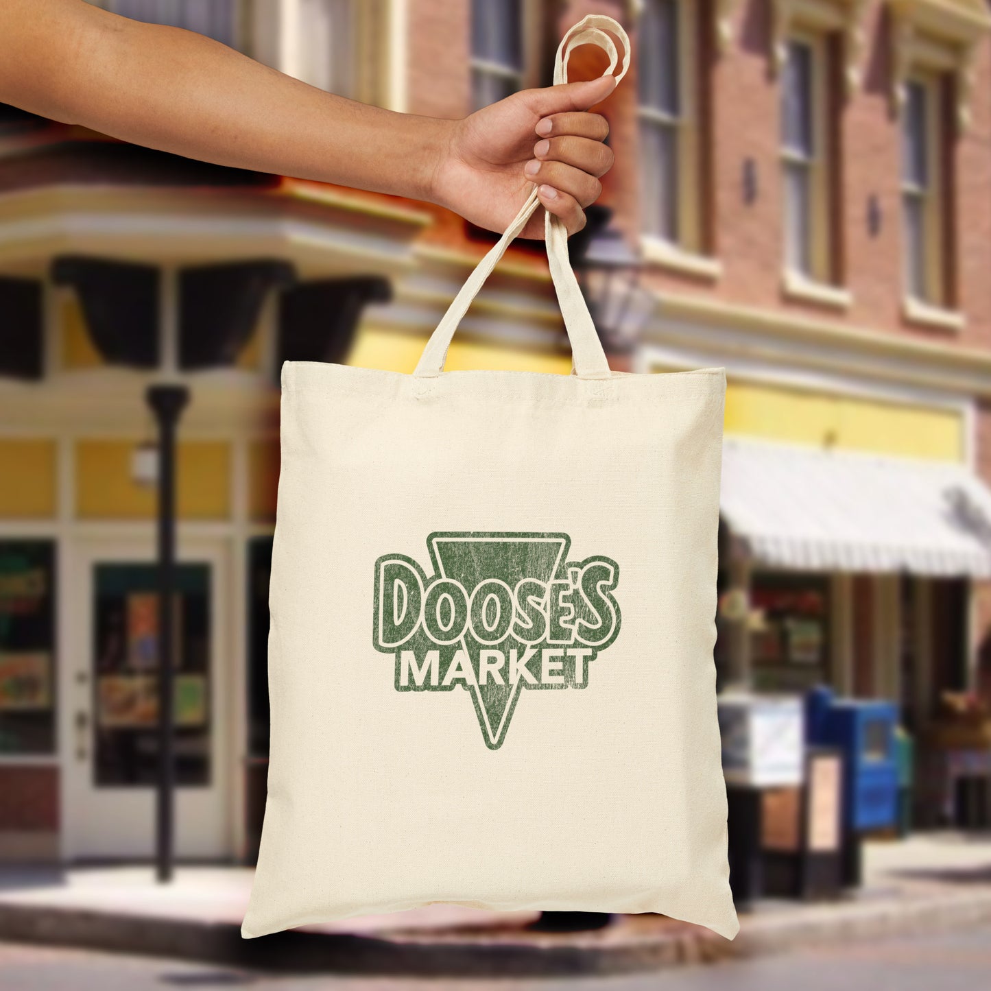 Person holding cotton canvas tote bag with vintage green Doose’s Market logo on both sides in front of store, inspired by Gilmore Girls.