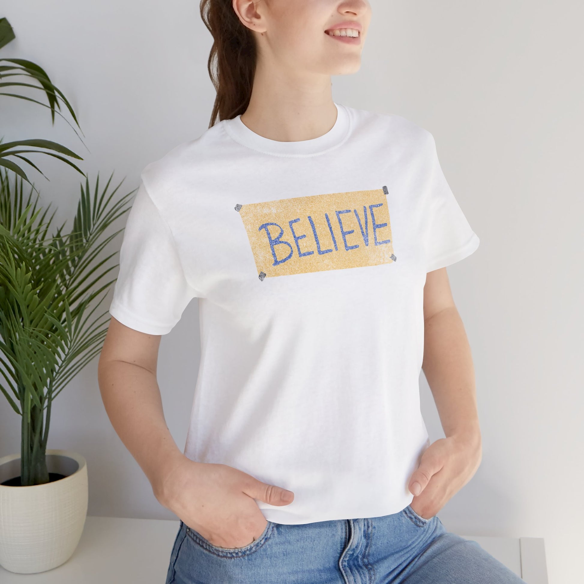 Female model wearing a white colored soft Bella+Canvas 3001 short-sleeve t-shirt featuring a logo graphic of the BELIEVE sign Ted Lasso hung in the fictional English soccer team AFC Richmond locker room, inspired by the TV show Ted Lasso in a vintage distressed style, sold by ScriptedBrand.