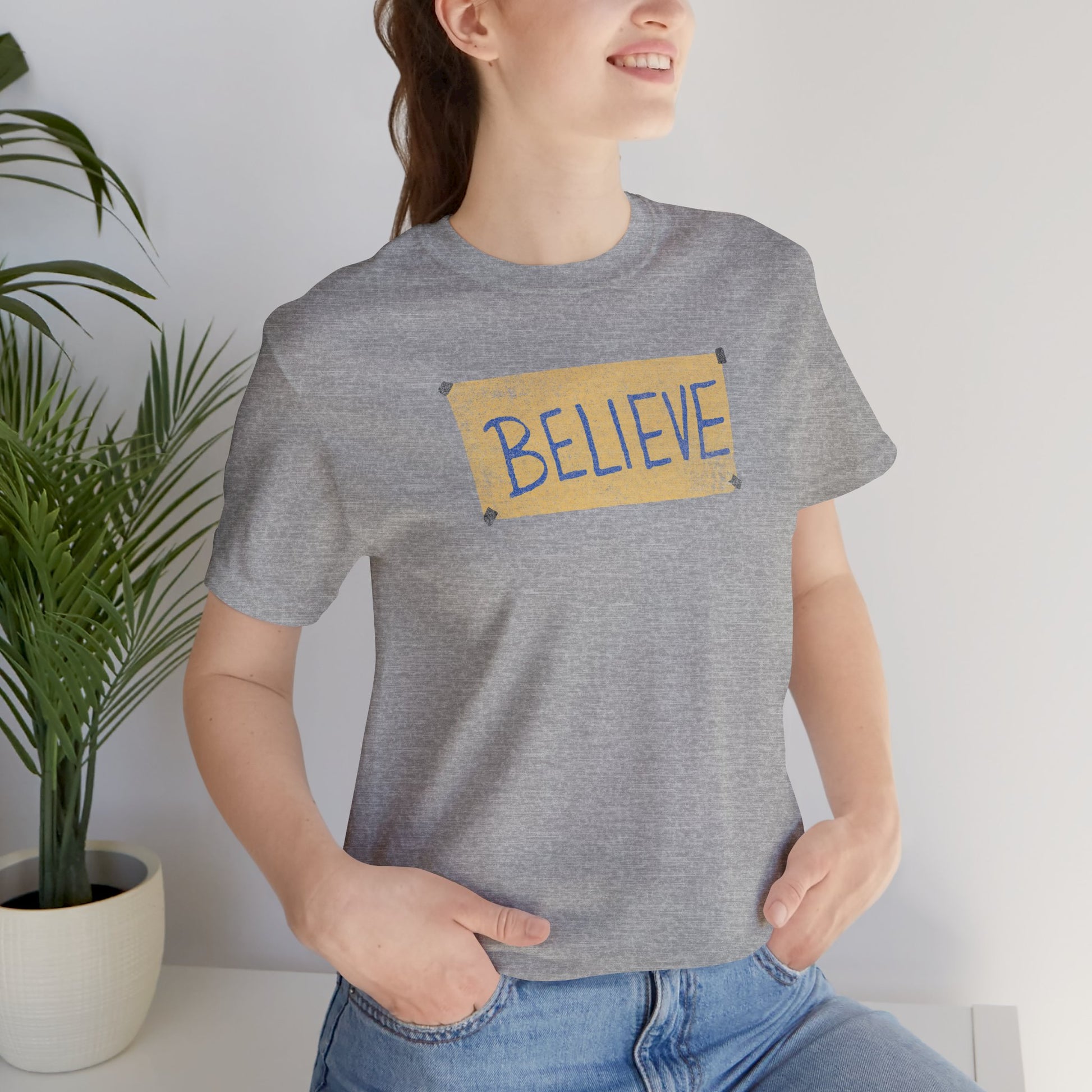 Female model wearing an athletic heather grey colored soft Bella+Canvas 3001 short-sleeve t-shirt featuring a logo graphic of the BELIEVE sign Ted Lasso hung in the fictional English soccer team AFC Richmond locker room, inspired by the TV show Ted Lasso in a vintage distressed style, sold by ScriptedBrand.