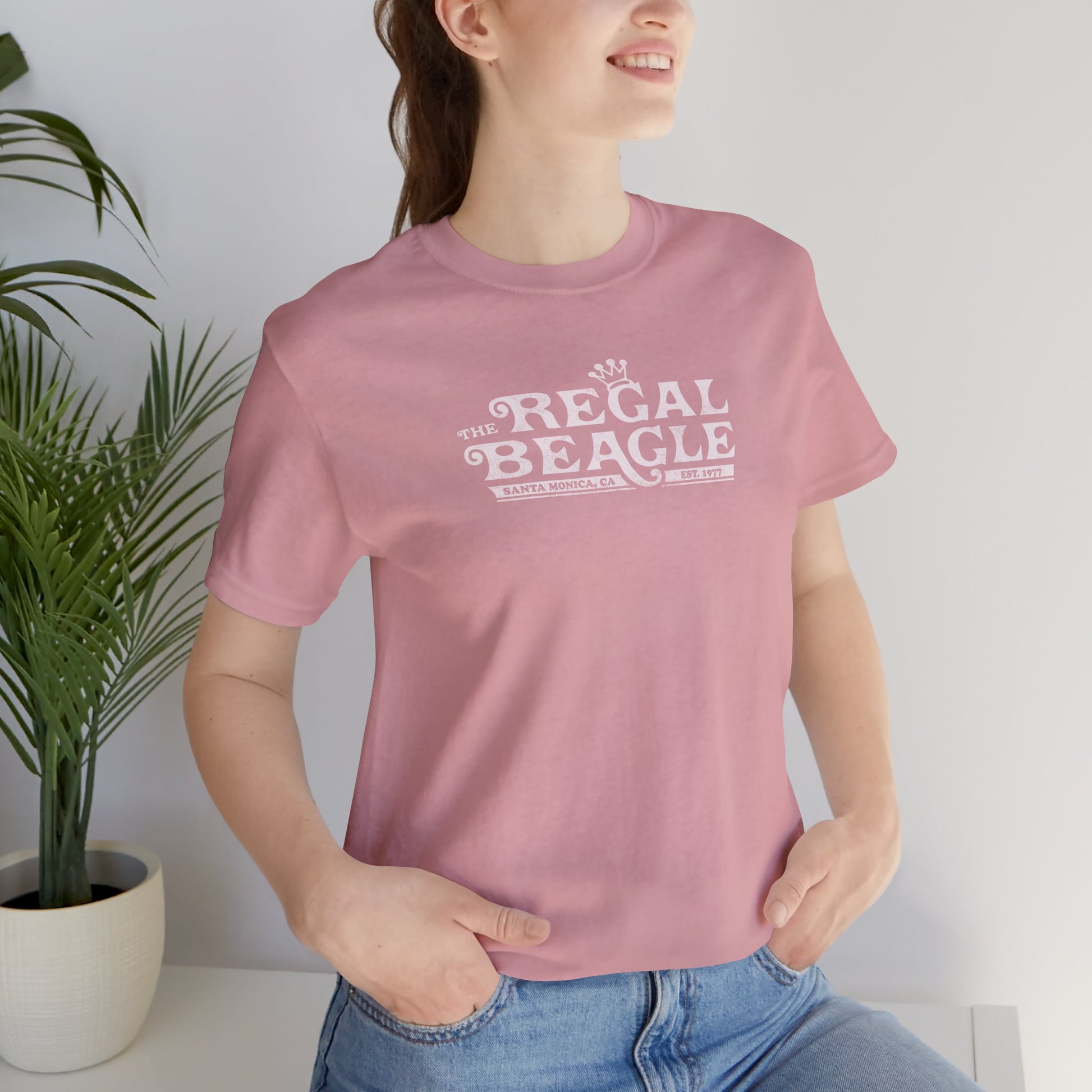 Female model wearing an orchid pink colored soft Bella+Canvas 3001 short-sleeve t-shirt featuring the Regal Beagle logo from the TV Show Threes Company in a vintage distressed style.
