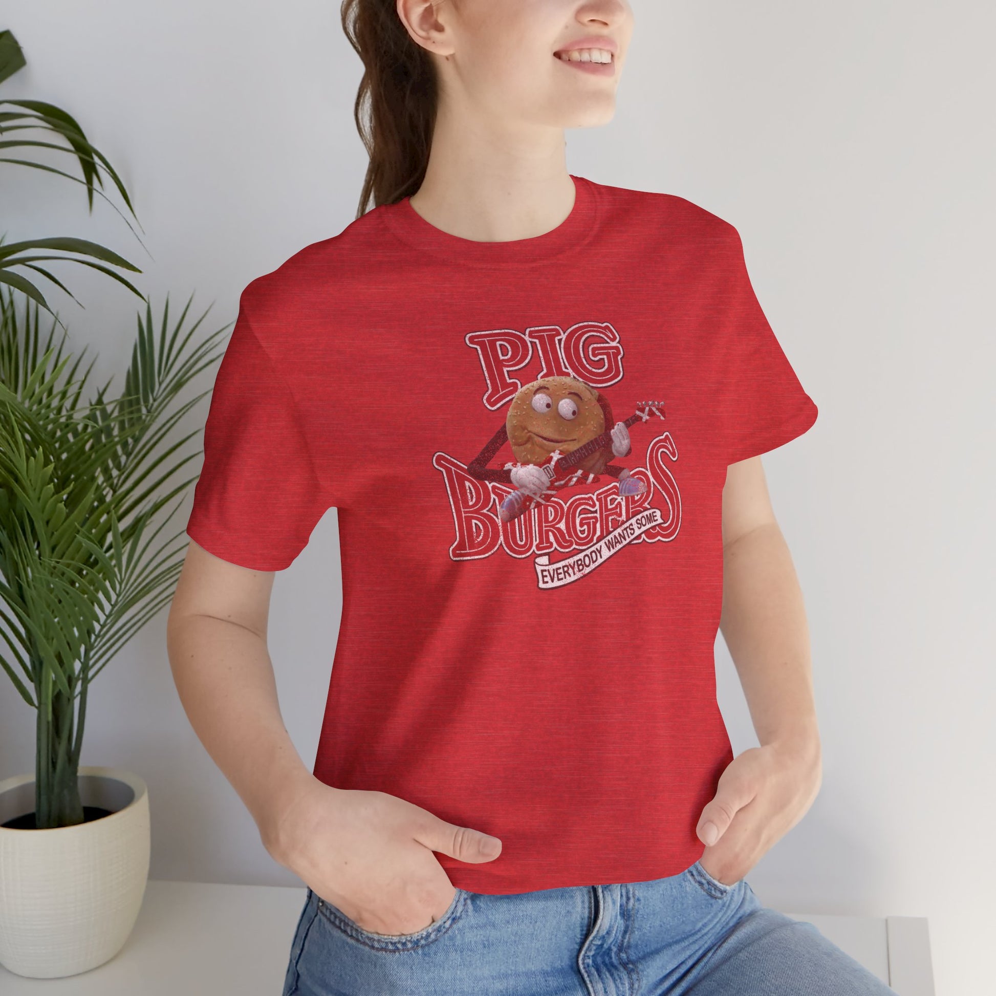 Female model wearing a heather red colored soft Bella+Canvas 3001 short-sleeve t-shirt featuring a graphic of the fictional Pig Burgers logo from the movie Better Off Dead, along with the Van Halen singing hamburger playing guitar, with the small subhead phrase EVERYBODY WANTS SOME. The graphic is designed in a vintage distressed style, sold by ScriptedBrand.