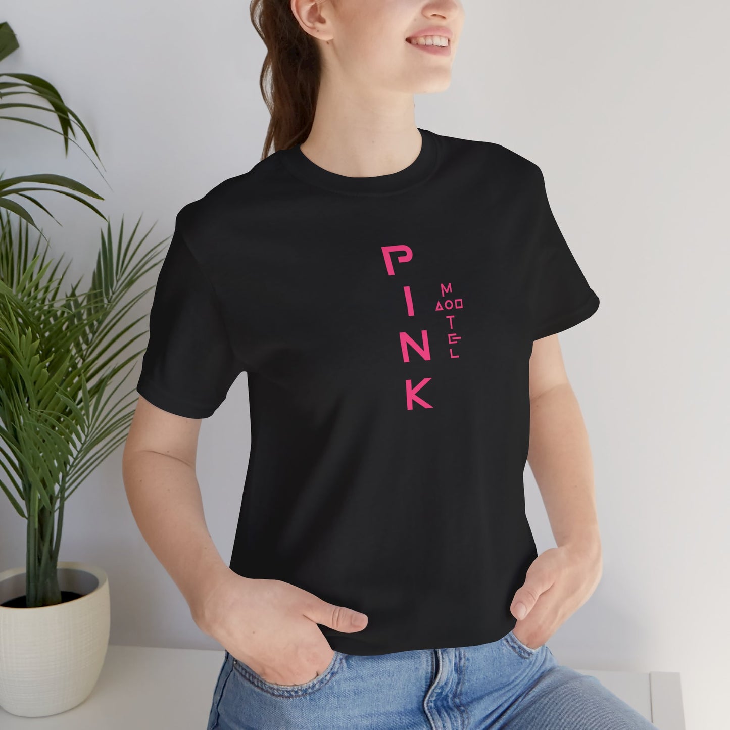 Female model wearing a black colored soft Bella+Canvas 3001 short-sleeve t-shirt featuring a pink logo graphic of the fictional Pink Motel inspired by the TV series Squid Game, in a vintage distressed style, sold by ScriptedBrand.