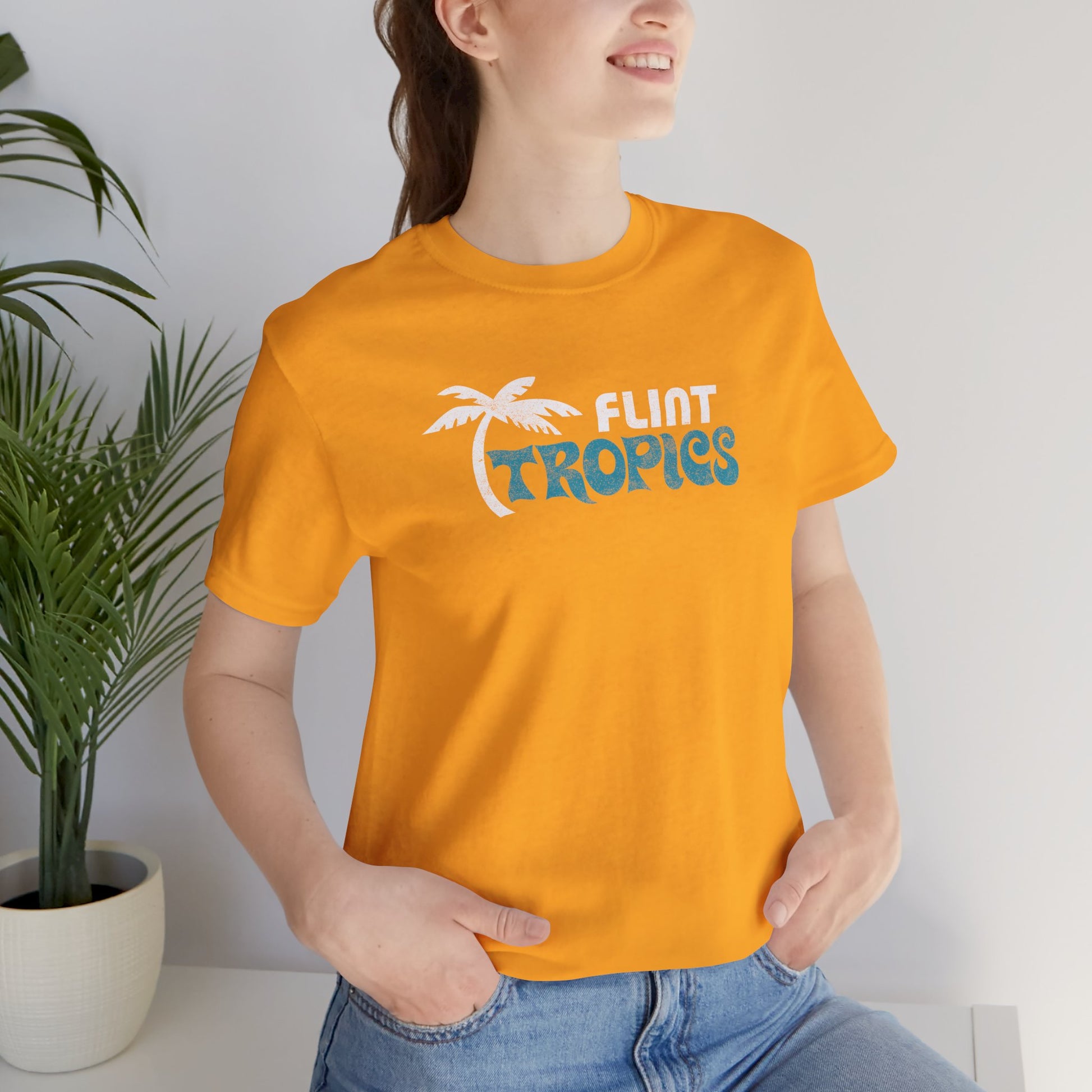 Female model wearing a gold colored soft Bella+Canvas 3001 short-sleeve t-shirt tee featuring the Flint Tropics logo from the movie Semi-Pro, in a vintage distressed style, sold by ScriptedBrand.