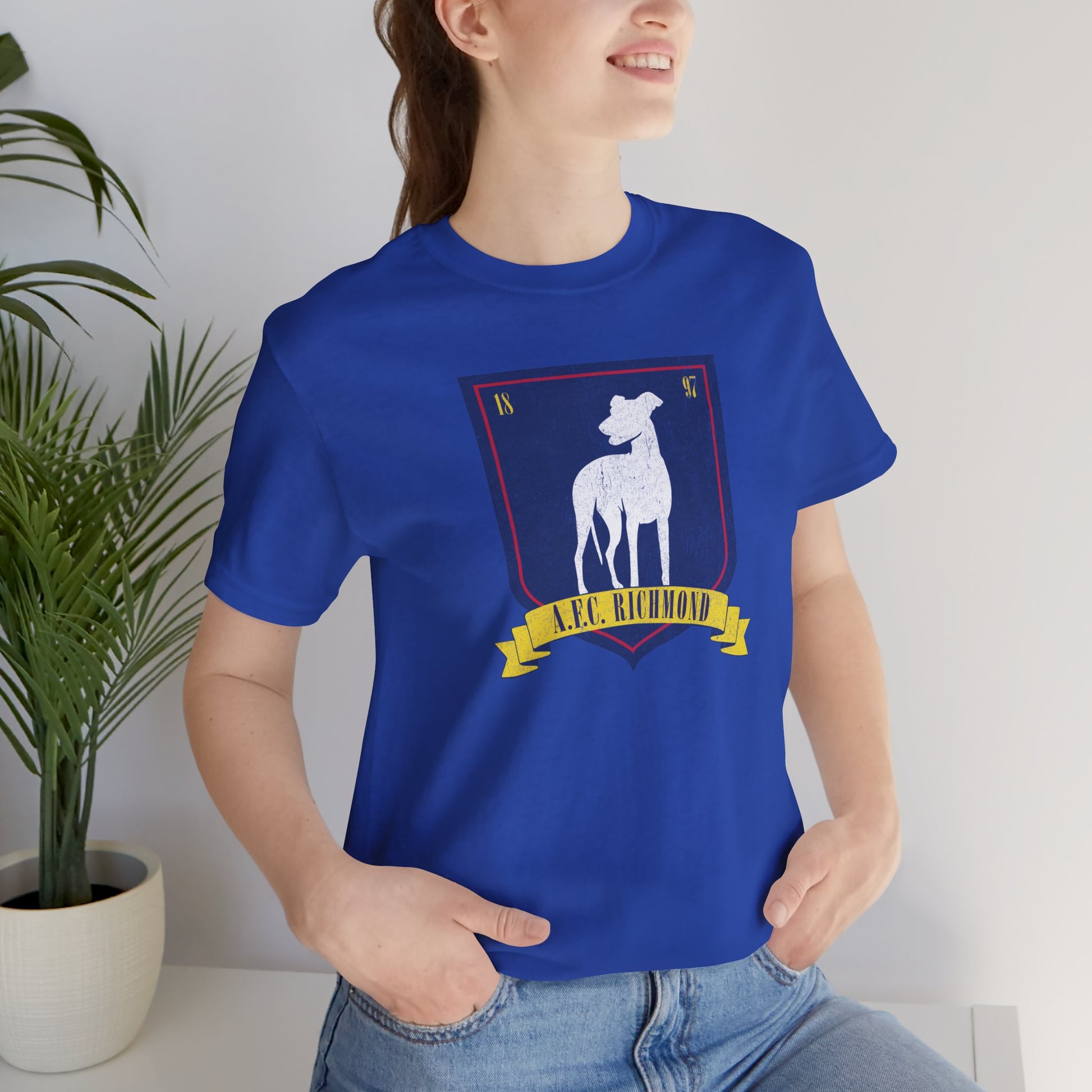 Female model wearing a royal blue soft Bella+Canvas 3001 short-sleeve t-shirt featuring the logo graphic of the fictional English soccer team AFC Richmond inspired by the TV show Ted Lasso in a vintage distressed style, sold by ScriptedBrand.