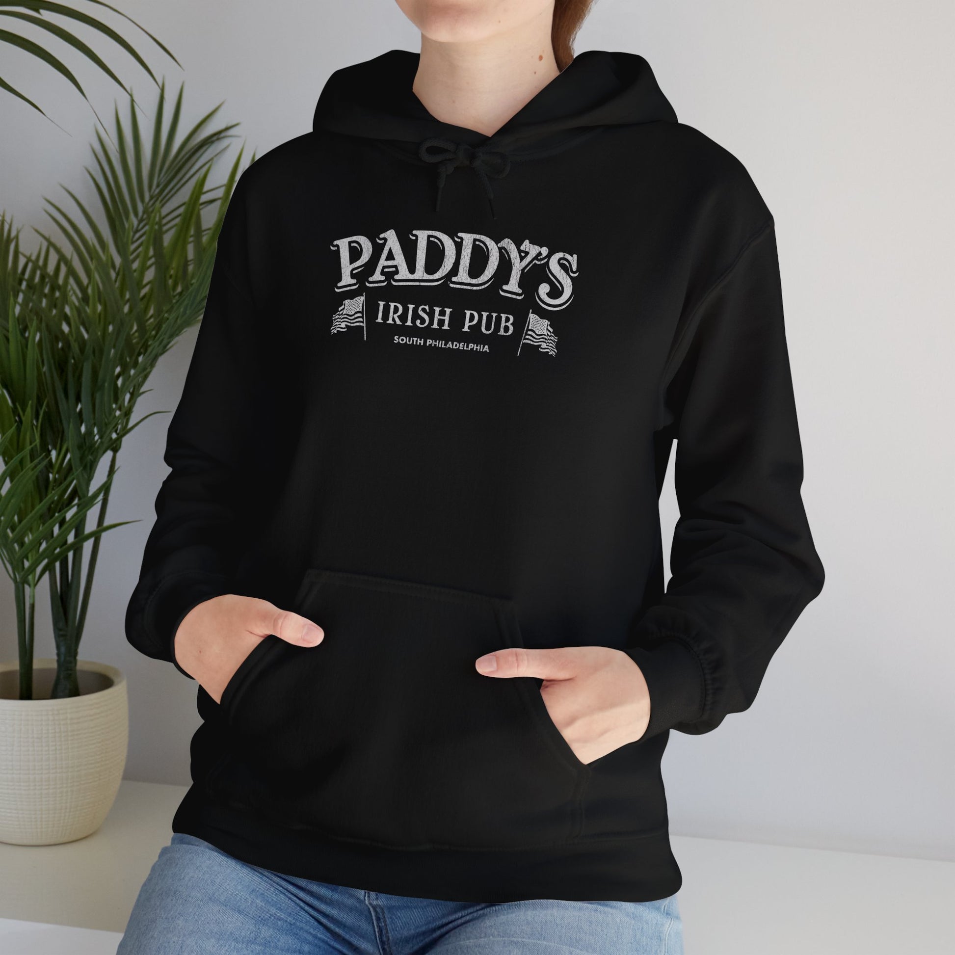 Female model wearing a black colored soft unisex Hoodie Heavy Blend Hooded Sweatshirt featuring the Paddy's Pub logo graphic from the TV Show It's Always Sunny in Philadelphia, in a vintage distressed style, sold by ScriptedBrand.