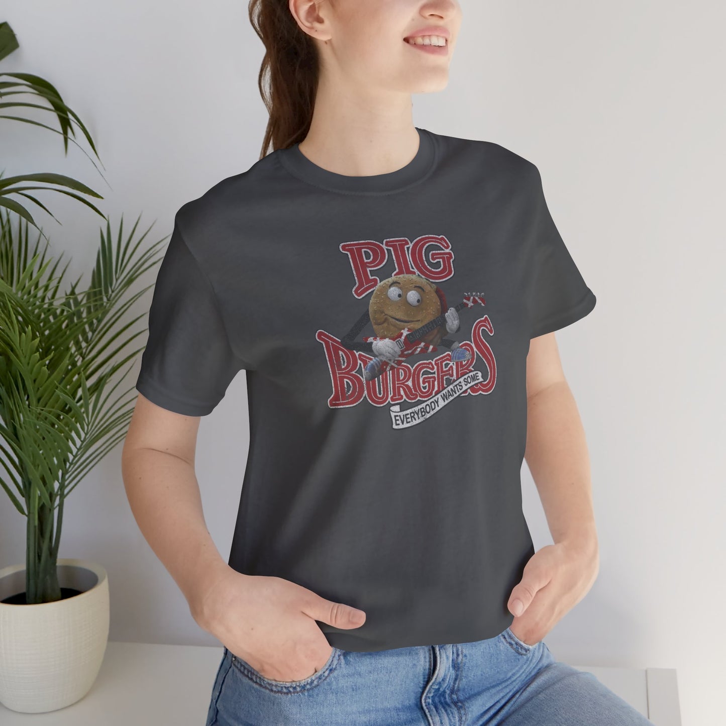 Female model wearing an asphalt grey colored soft Bella+Canvas 3001 short-sleeve t-shirt featuring a graphic of the fictional Pig Burgers logo from the movie Better Off Dead, along with the Van Halen singing hamburger playing guitar, with the small subhead phrase EVERYBODY WANTS SOME. The graphic is designed in a vintage distressed style, sold by ScriptedBrand.