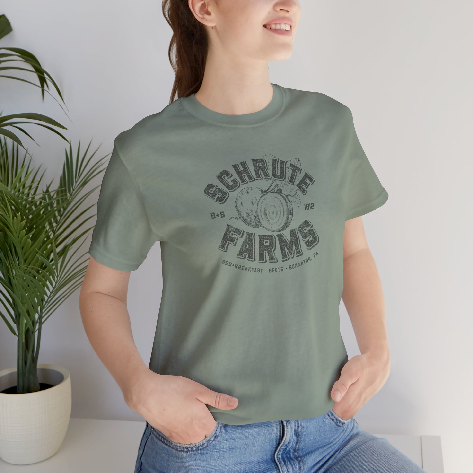 Person wearing a Sage green t-shirt featuring a Schrute Farms logo from the TV show The Office designed by ScriptedBrand in a vintage distressed style.