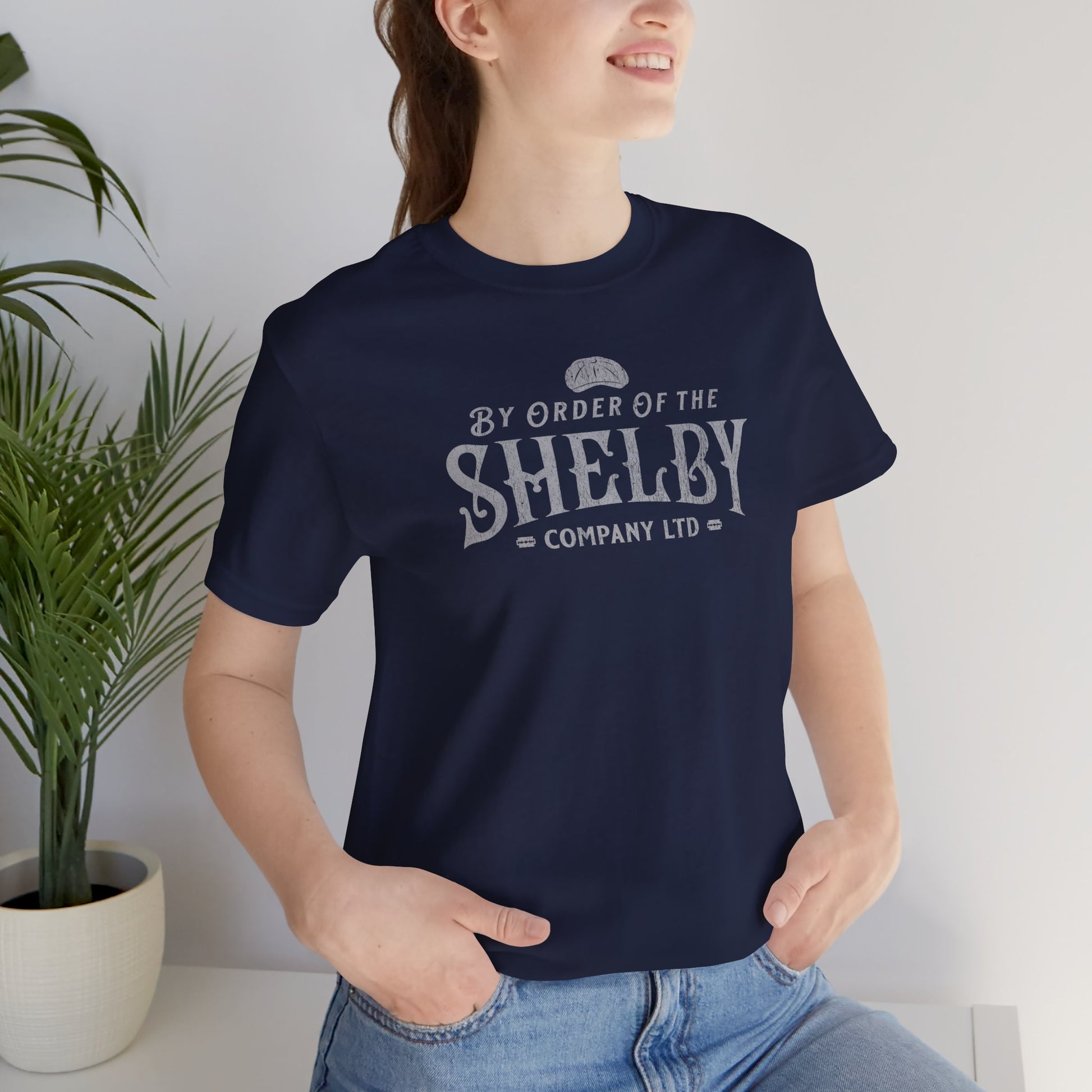 Female model wearing a navy blue grey colored soft short-sleeve Bella+Canvas 3001 t-shirt featuring a unique Shelby Company Limited logo graphic from the show Peaky Blinders and movie The Immortal Man, in a vintage distressed style, includes a flat cap and razor blades, designed and sold by ScriptedBrand.