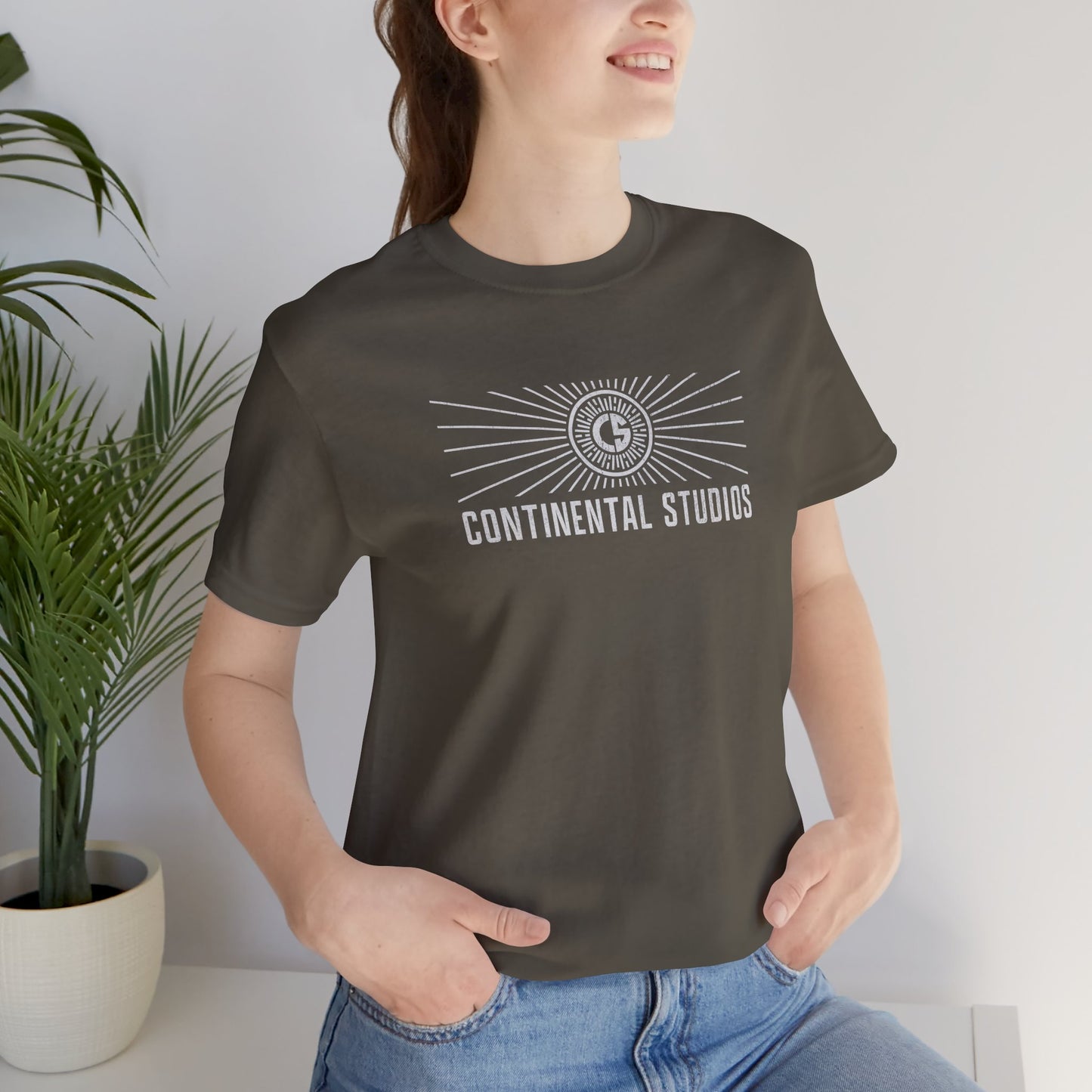 Female model wearing an army green colored Soft Bella+Canvas 3001 short-sleeve t-shirt featuring the logo graphic of the fictional movie studio Continental Studios inspired by the Apple TV show The Studio in a vintage distressed style, sold by ScriptedBrand.