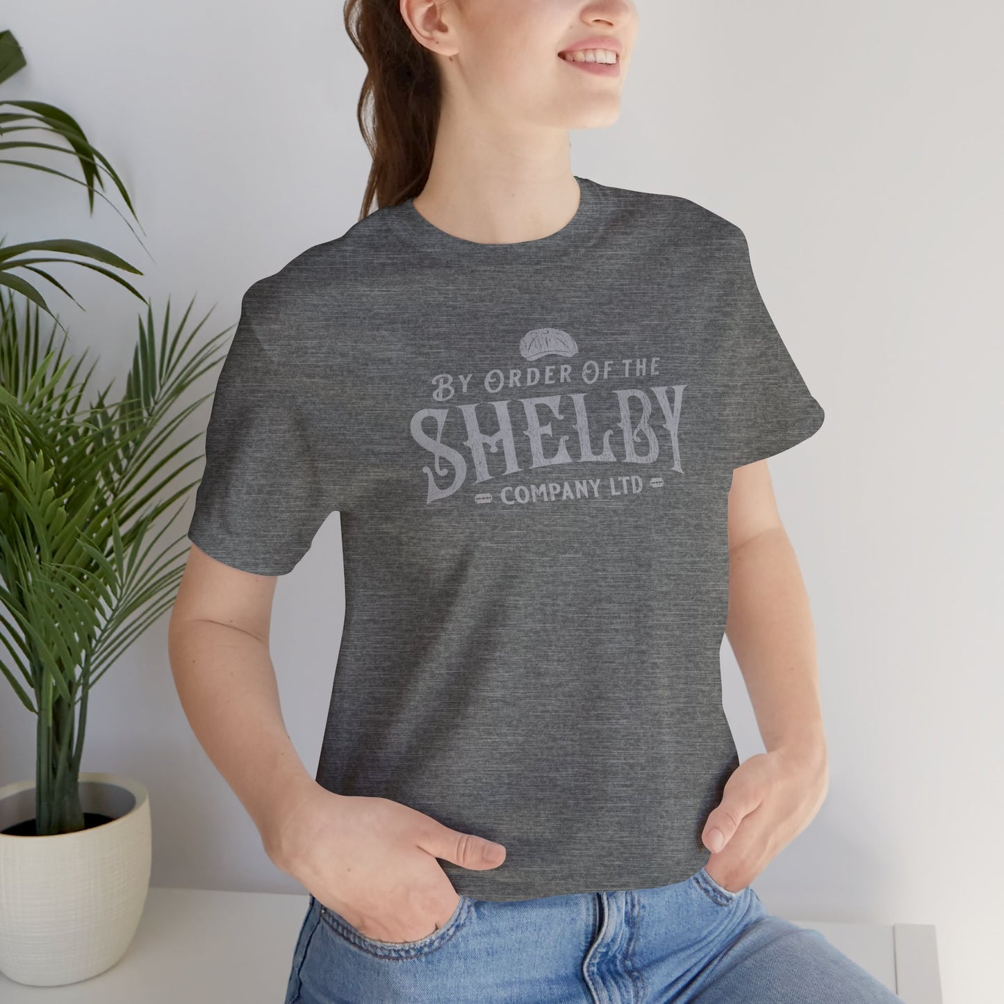Female model wearing a deep heather grey colored soft short-sleeve Bella+Canvas 3001 t-shirt featuring a unique Shelby Company Limited logo graphic from the show Peaky Blinders and movie The Immortal Man, in a vintage distressed style, includes a flat cap and razor blades, designed and sold by ScriptedBrand.