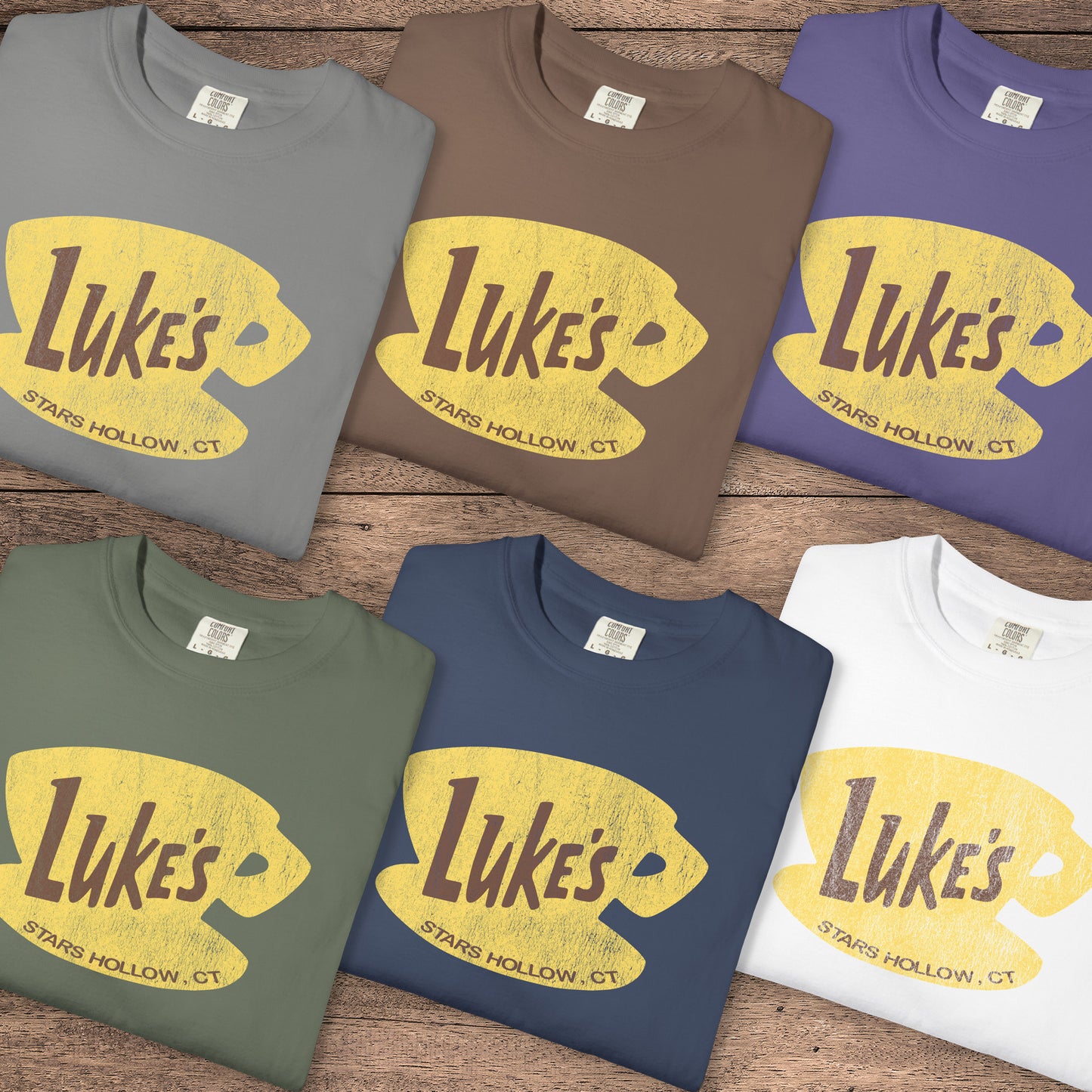 Six folded Comfort Colors t-shirts with vintage yellow Luke’s Diner logo from Gilmore Girls, laid flat on wood background.