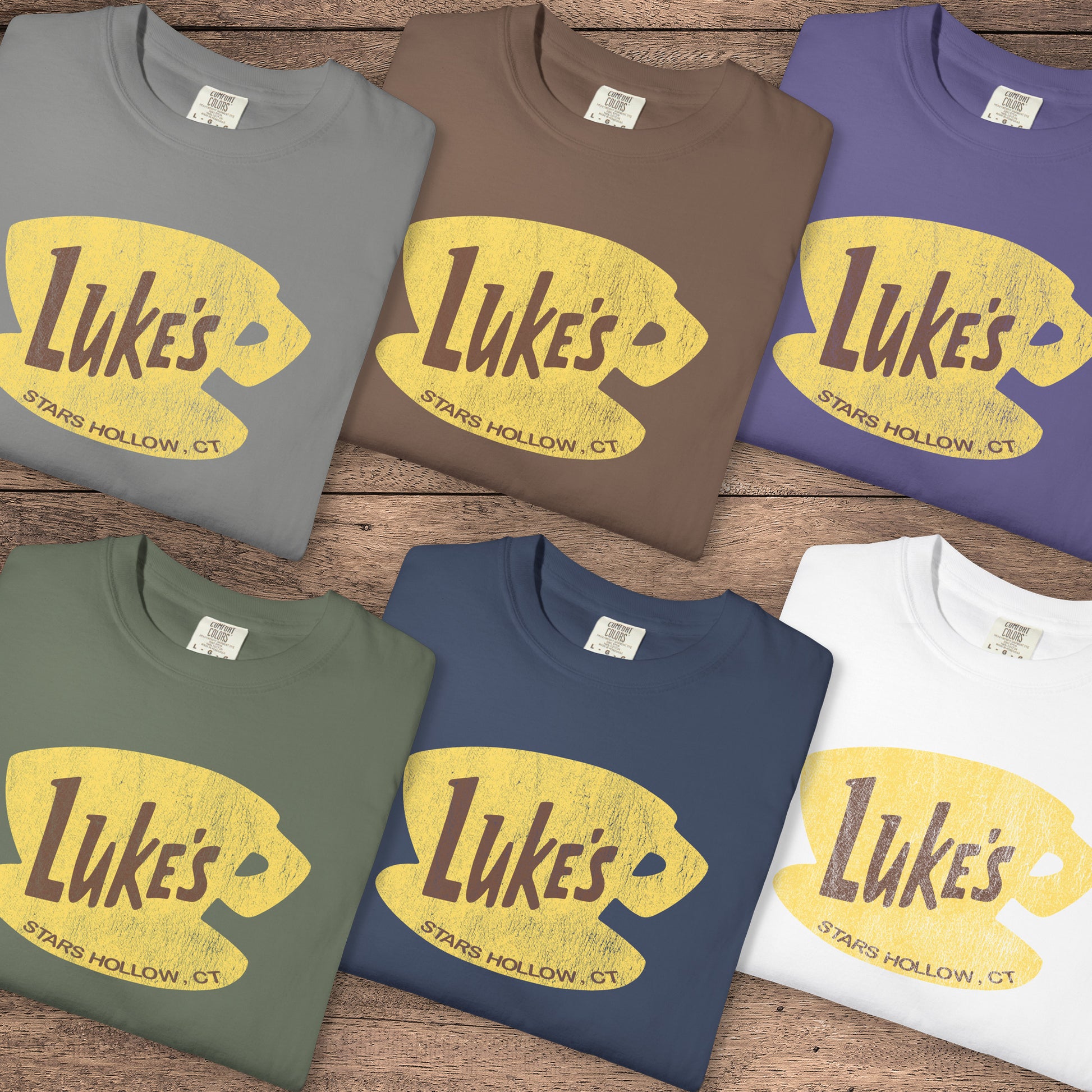 Six folded Comfort Colors t-shirts with vintage yellow Luke’s Diner logo from Gilmore Girls, laid flat on wood background.